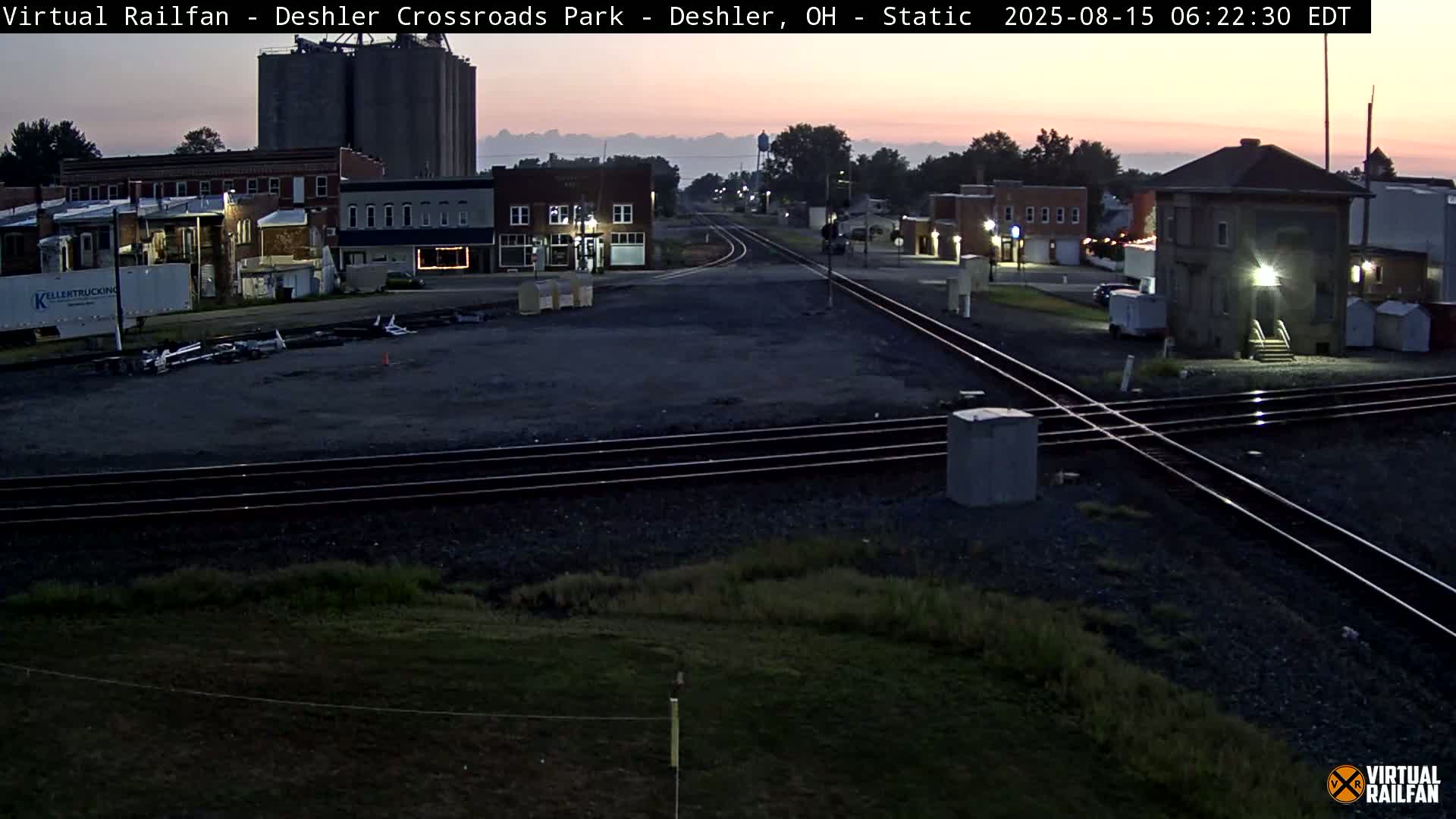 A nighttime view of a railroad crossing in a small town, with several buildings visible in the background.