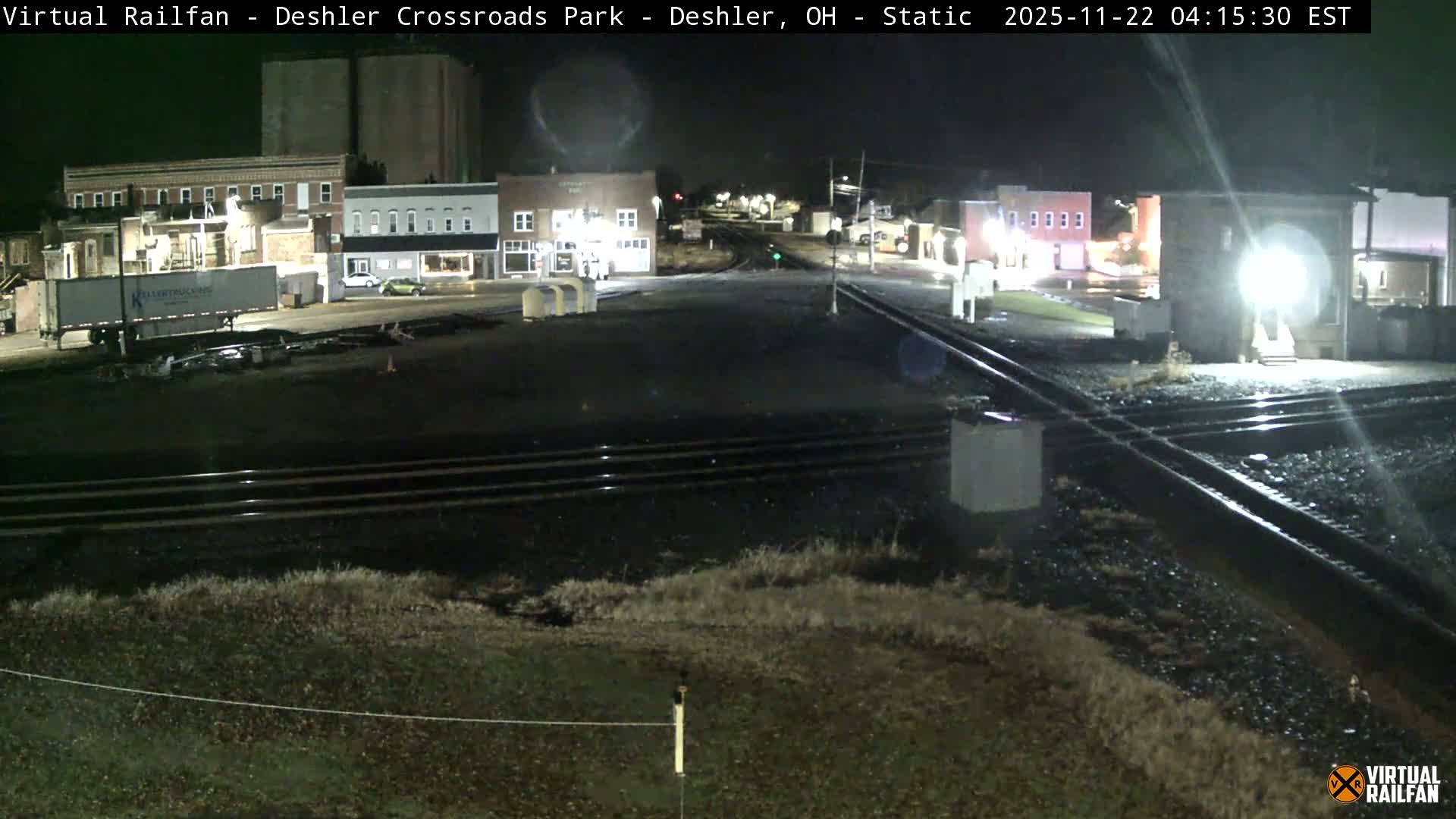Multiple railroad tracks dominate the foreground of an illuminated small town scene at night, featuring several brightly lit buildings, a semi-truck trailer, and streetlights under a clear sky.