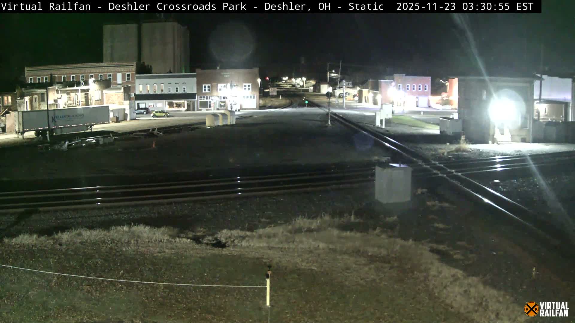 The image displays a clear night scene of a small town with several illuminated buildings, parked vehicles, and multiple train tracks in the foreground.