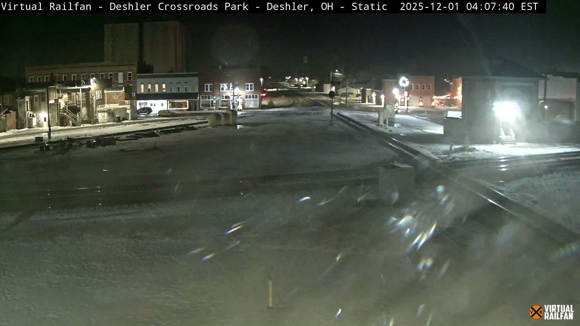 A dark winter night scene of a small town features snow-covered ground, illuminated buildings and streets, active railroad tracks, and visible falling snow reflecting light.