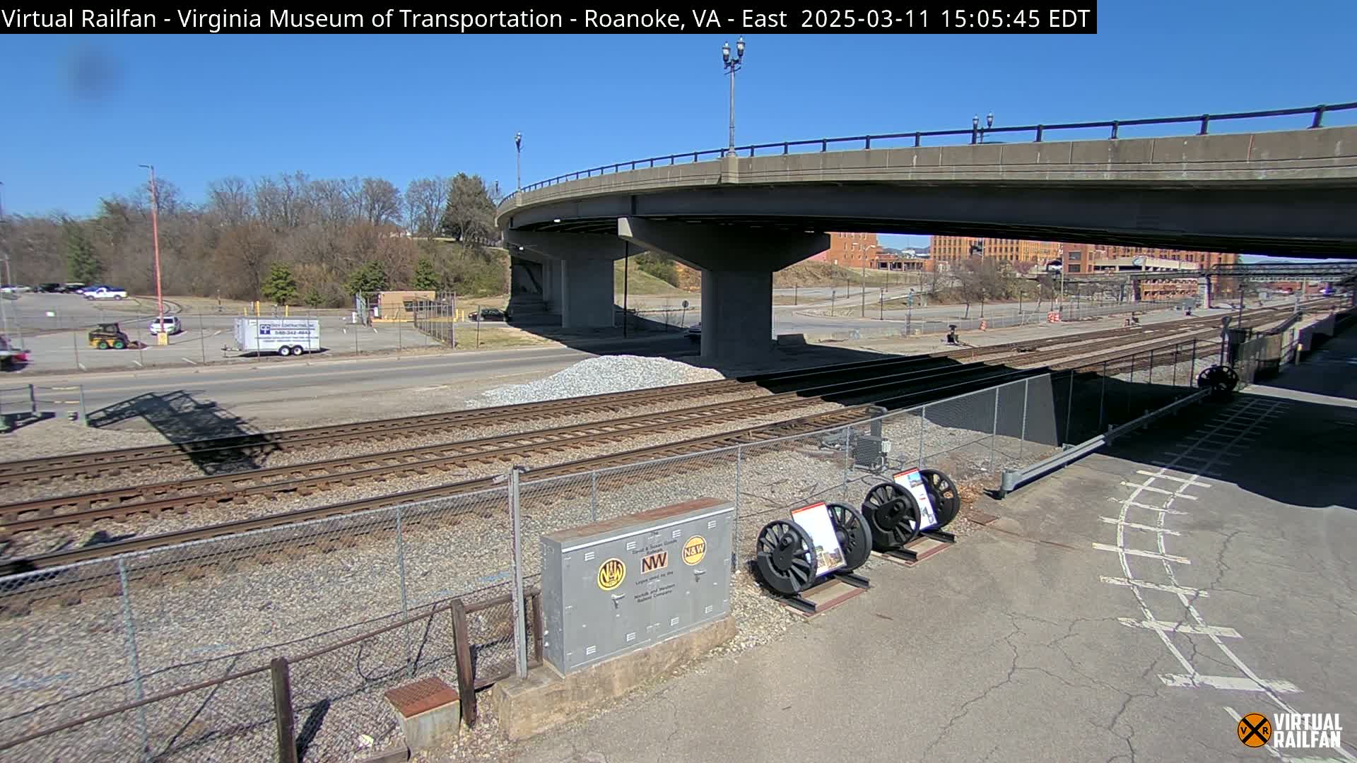 A sunny day shows railroad tracks, a roadway, a bridge, and some parked vehicles, with a pile of gravel nearby.