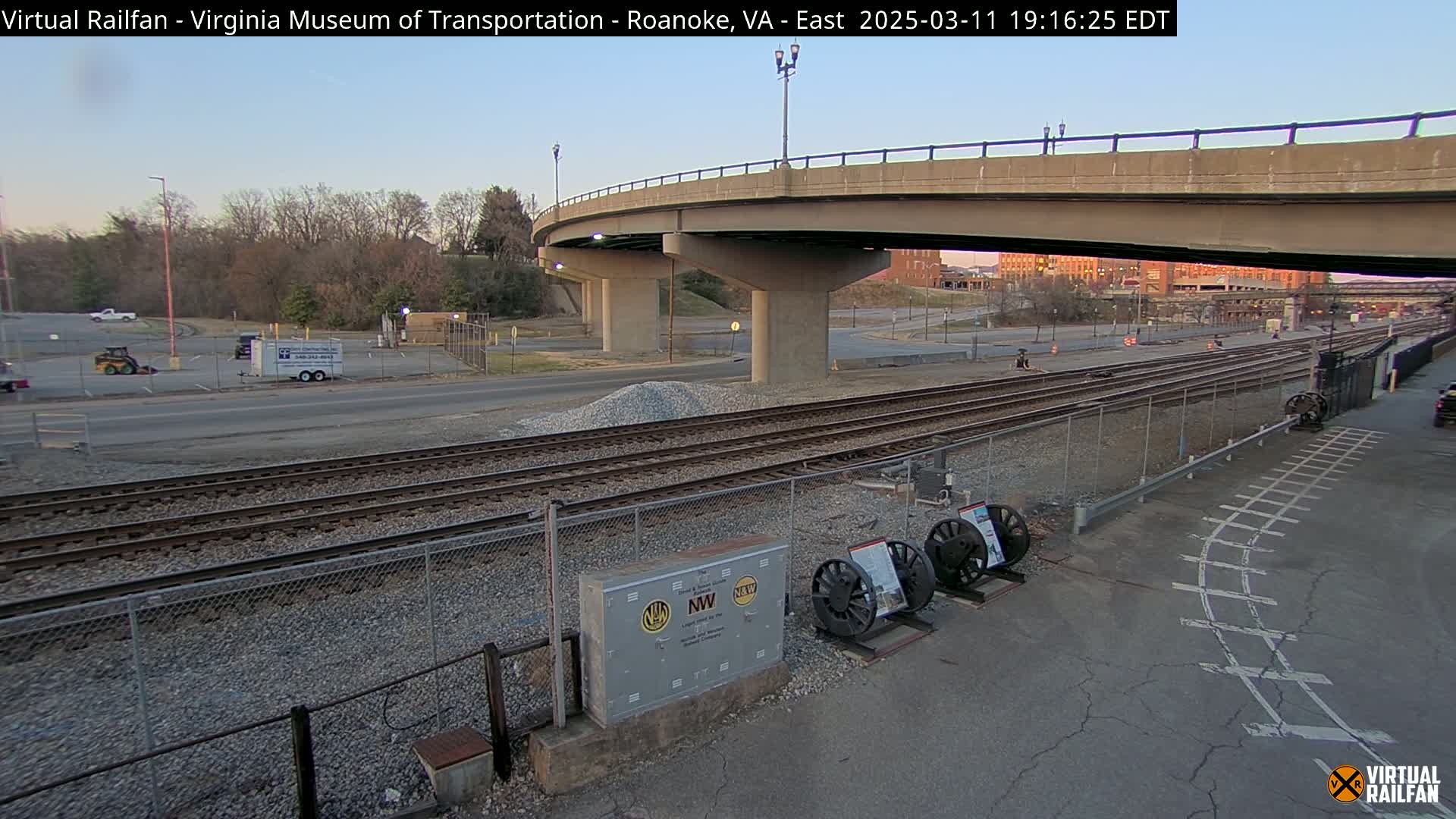 A twilight view of railroad tracks near a road and bridge, with a few vehicles and some construction materials visible.
