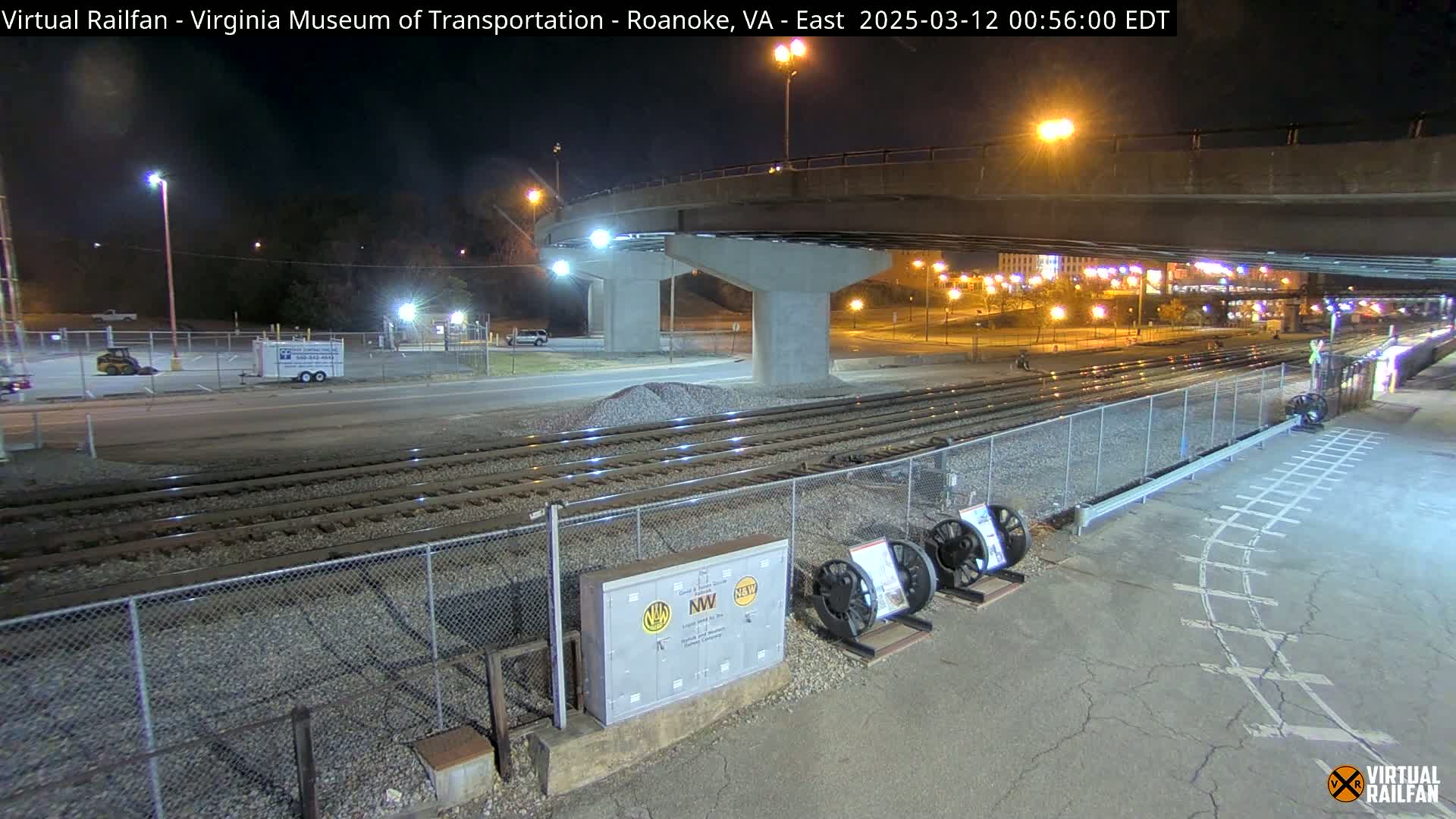 At night, train tracks run alongside a road under a bridge, with a few vehicles visible in a parking lot.