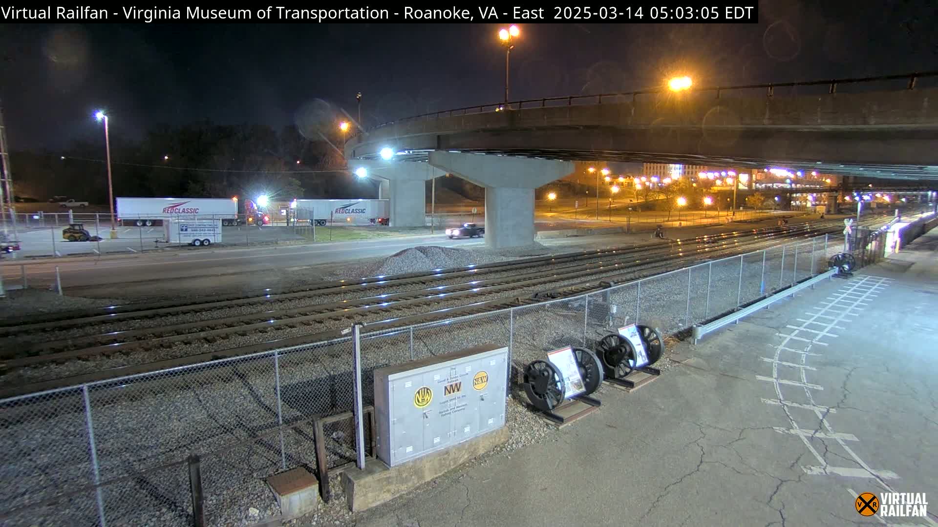 A nighttime view of railroad tracks under a bridge, with several semi-trucks and a car visible in the distance.