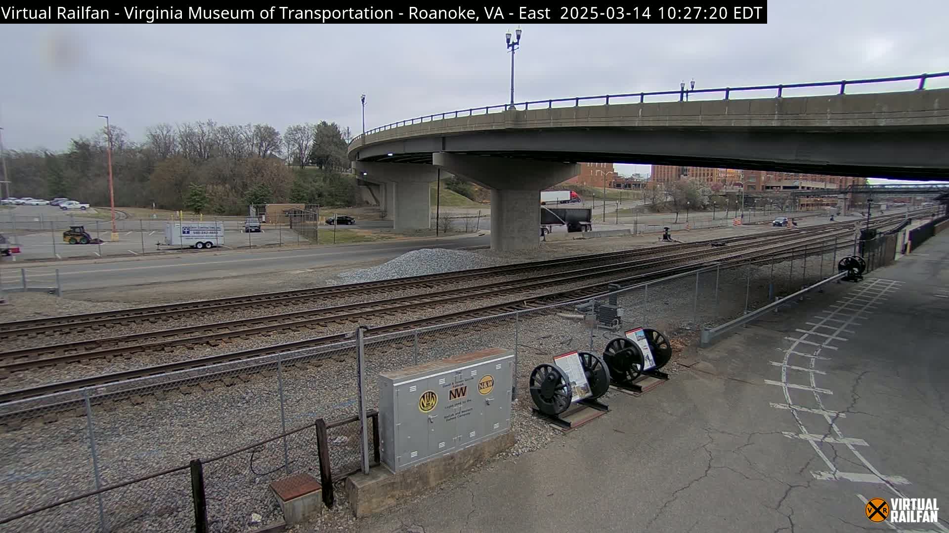 An overcast day reveals railroad tracks near a highway overpass, with a parking lot and some small vehicles visible in the background.