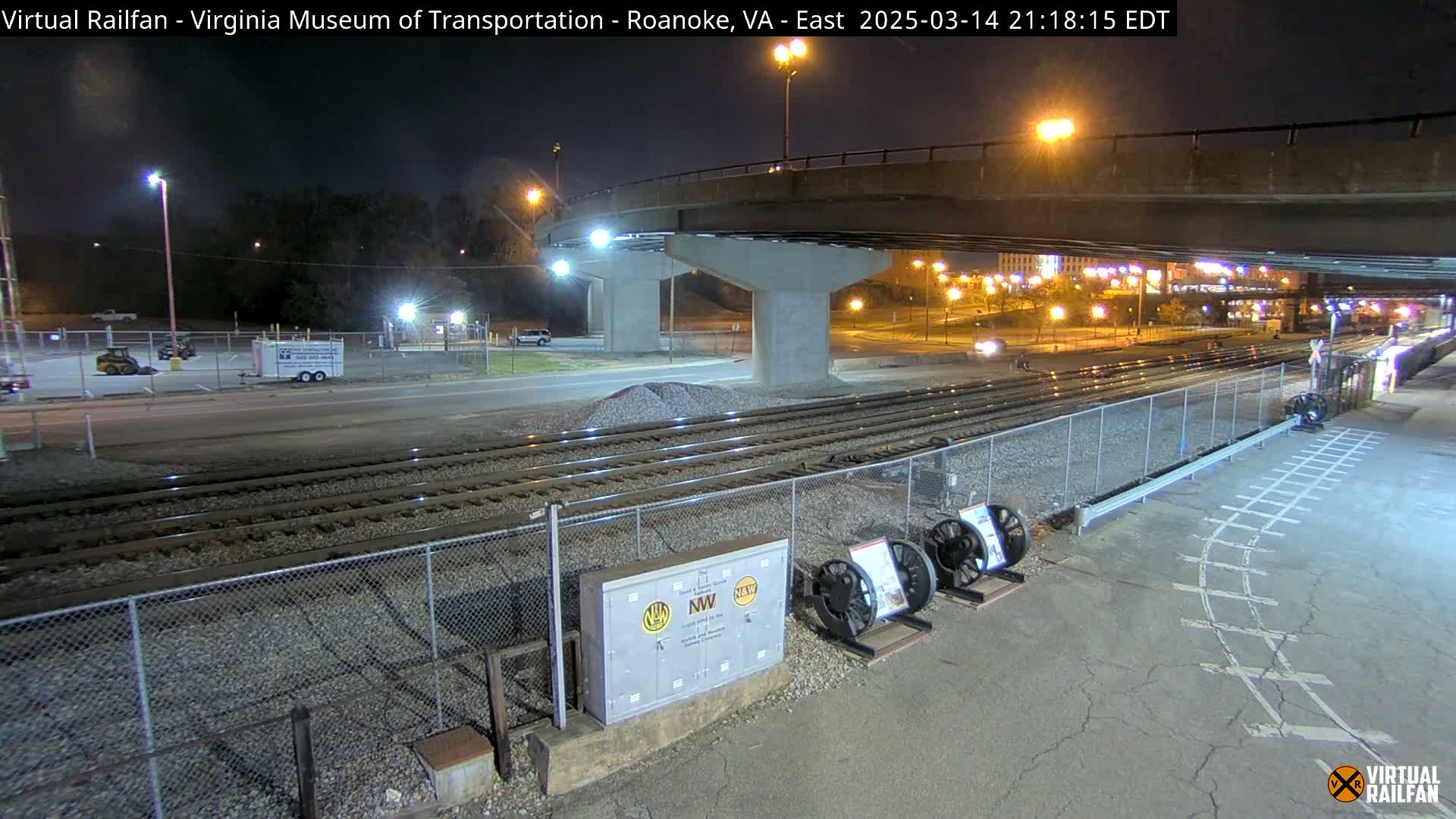A nighttime view of railroad tracks under a bridge, with a few vehicles visible in a parking lot and some city lights in the background.