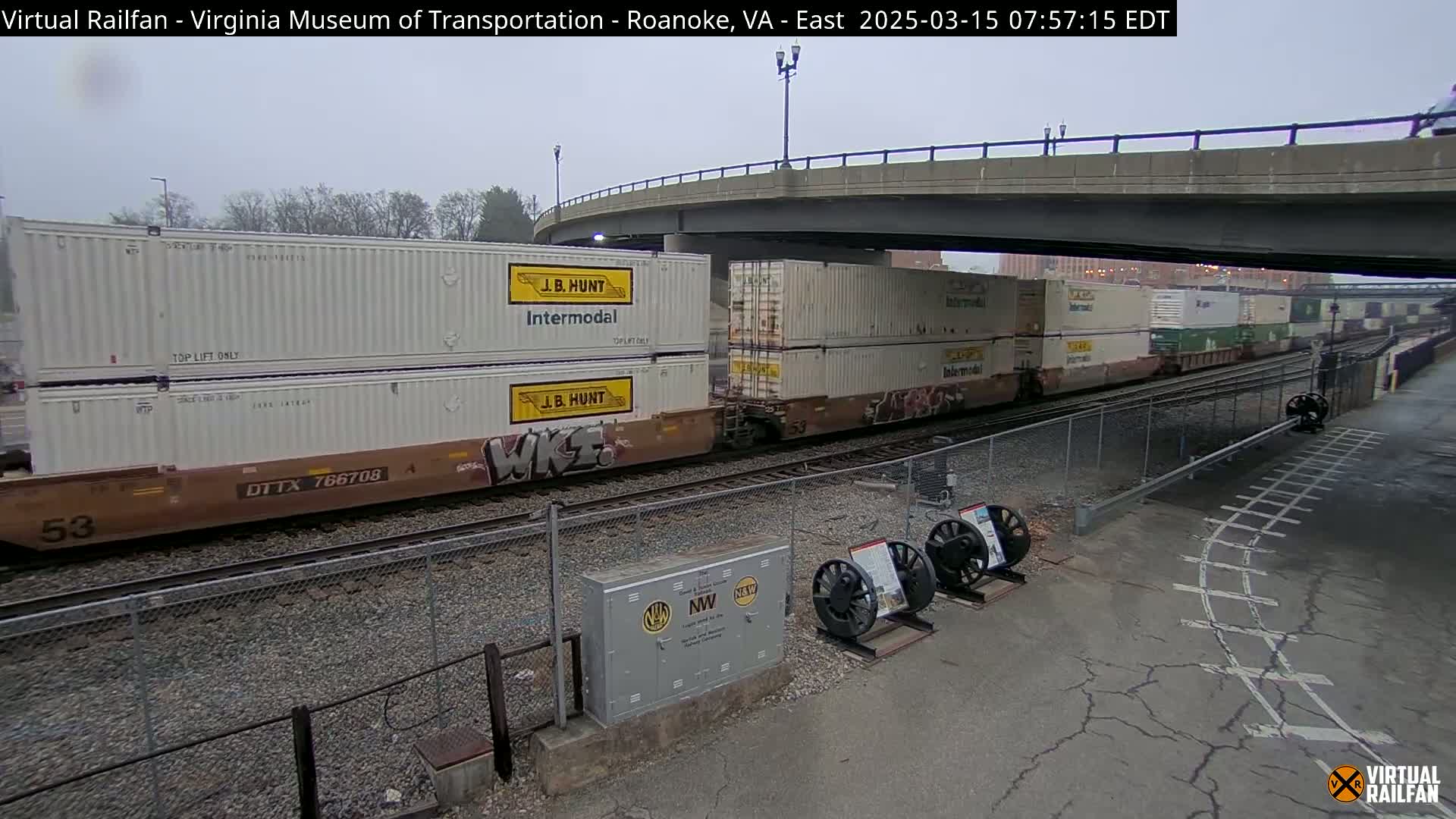 A long freight train carrying numerous shipping containers passes under an overpass on an overcast day.