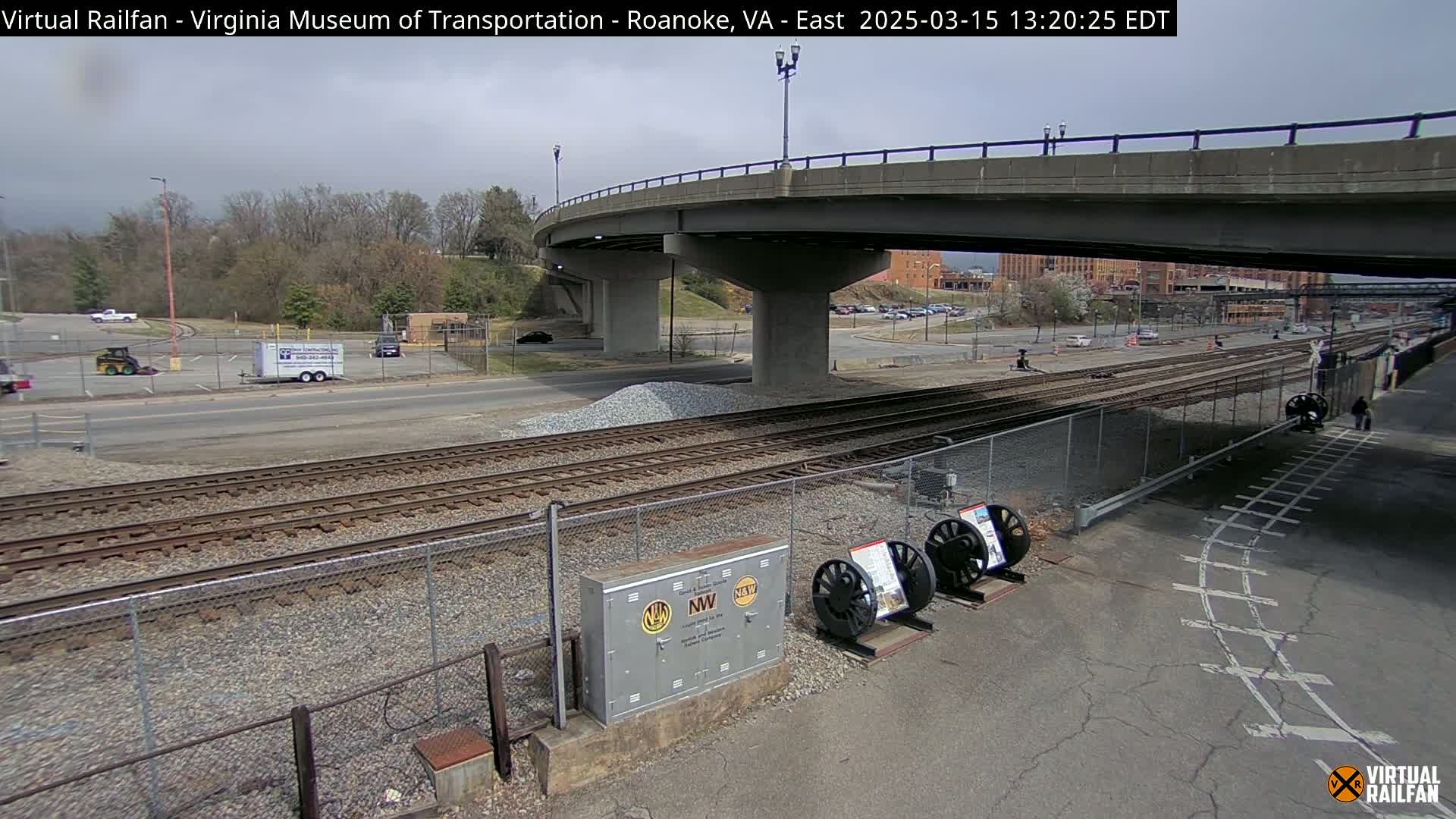 A mostly cloudy day reveals a view of railroad tracks, a roadway, and a bridge overpass, with some parked vehicles and construction materials visible.