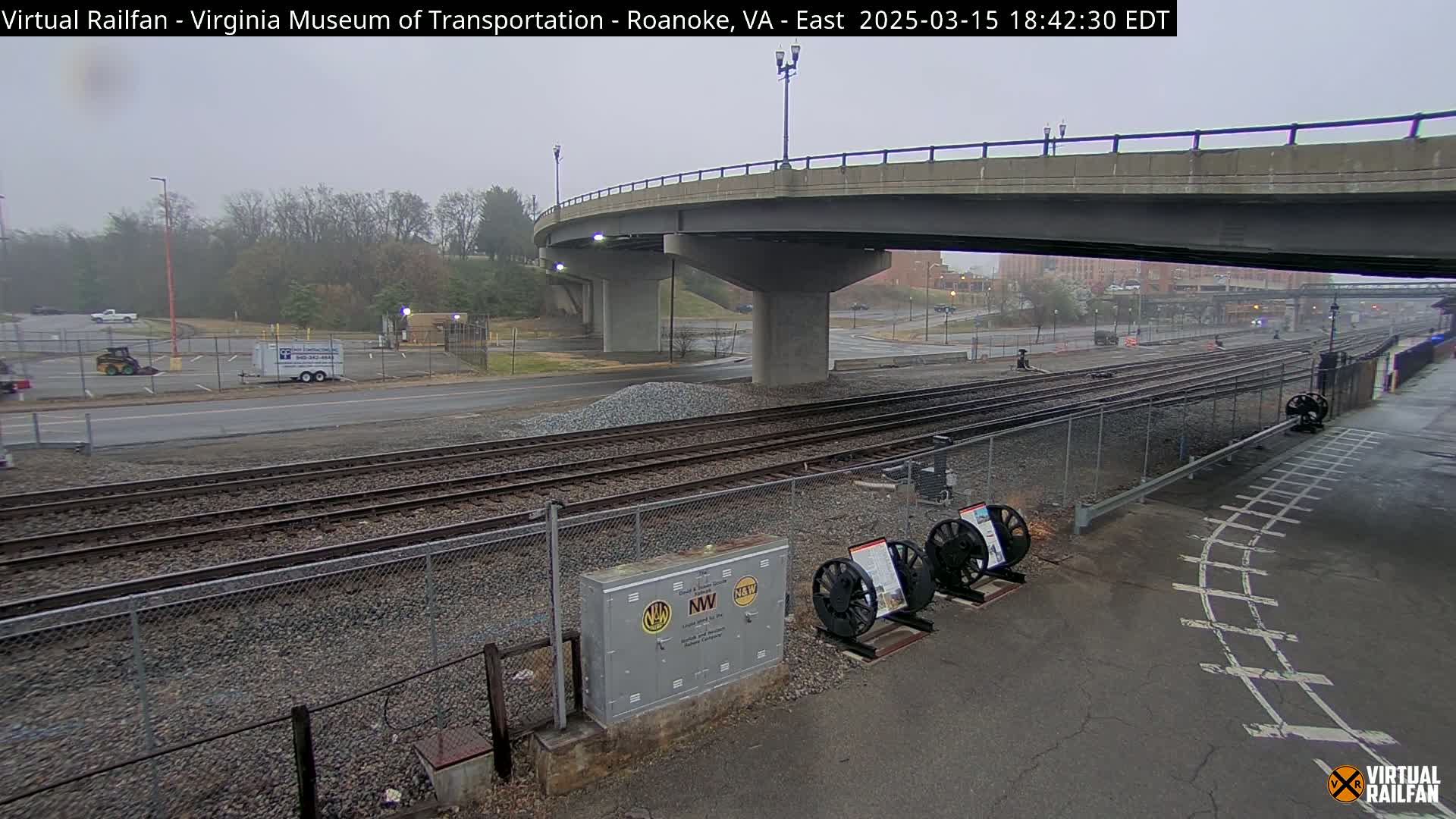 A mostly overcast day reveals railroad tracks and a roadway passing under a bridge, with some construction equipment and a small parking lot nearby.