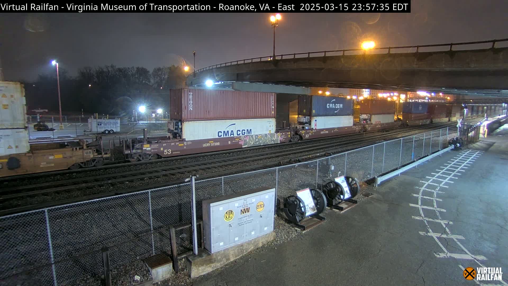 At night, under a bridge, a freight train carrying several shipping containers passes by a chain-link fence and some railway equipment.