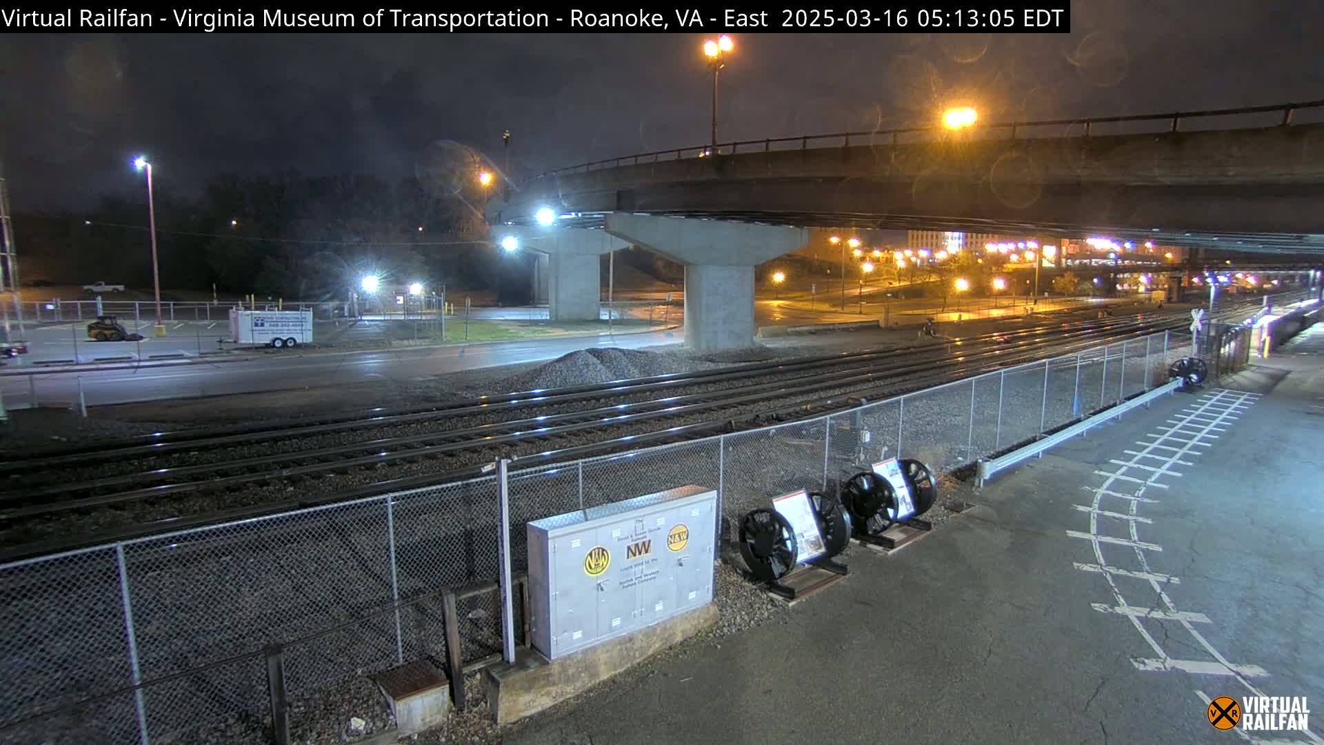 A nighttime view of train tracks beside a road under a bridge, with a parking lot and some vehicles visible in the background.