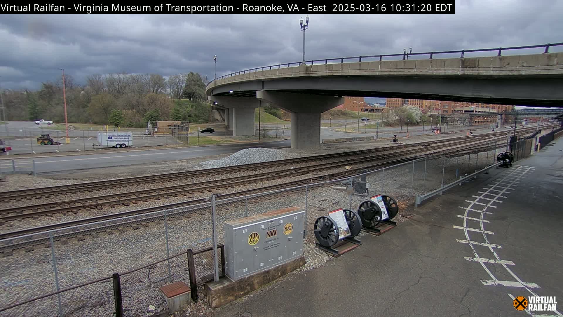 A cloudy day reveals railroad tracks extending under a bridge, with a small parking lot and construction materials nearby.