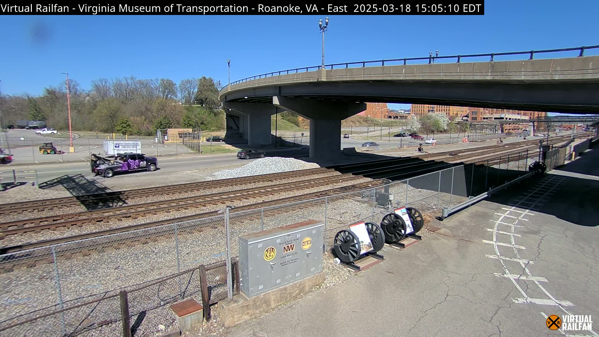 Under a sunny sky, railroad tracks run alongside a road beneath a bridge, with a purple tow truck and a pile of gravel visible.