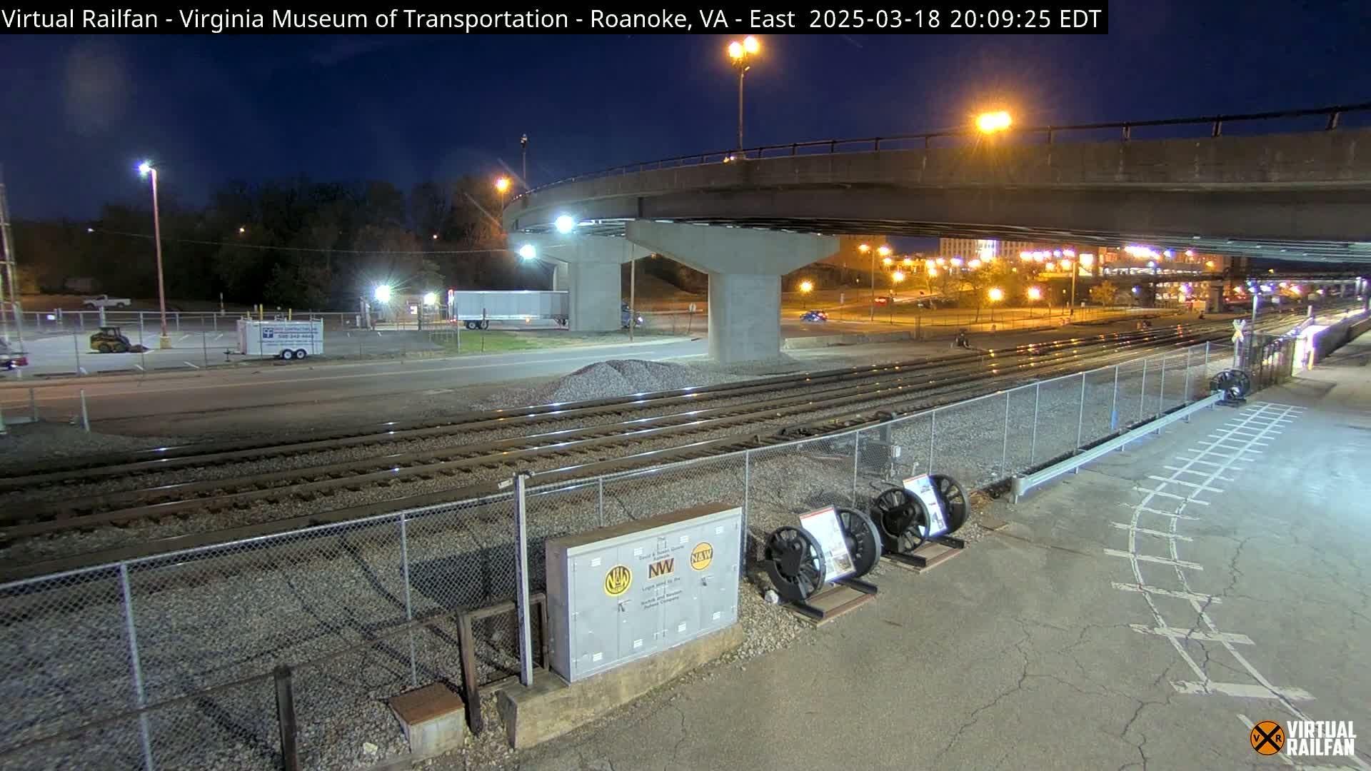 Under a dark night sky, railroad tracks extend beneath a highway overpass, with several pieces of railway equipment visible nearby.
