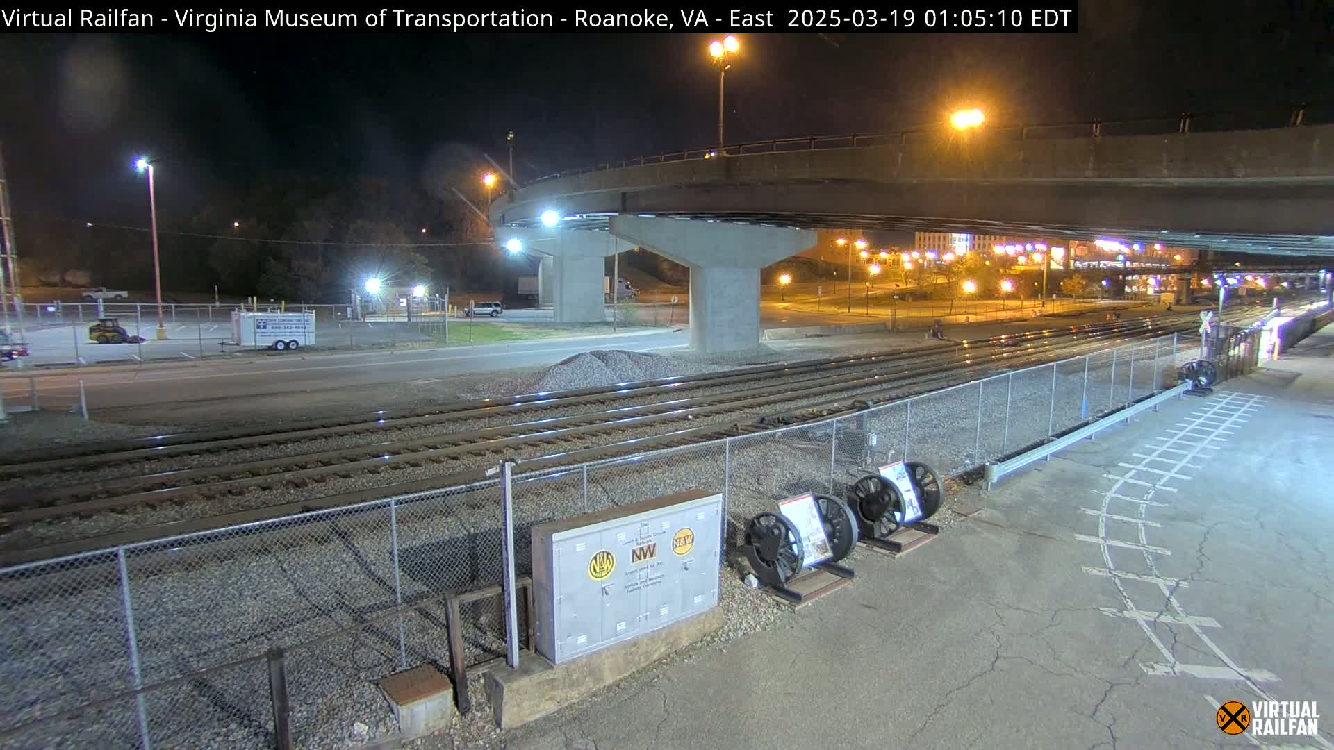 A nighttime view of train tracks next to a road under a bridge, with several cars visible in the distance.