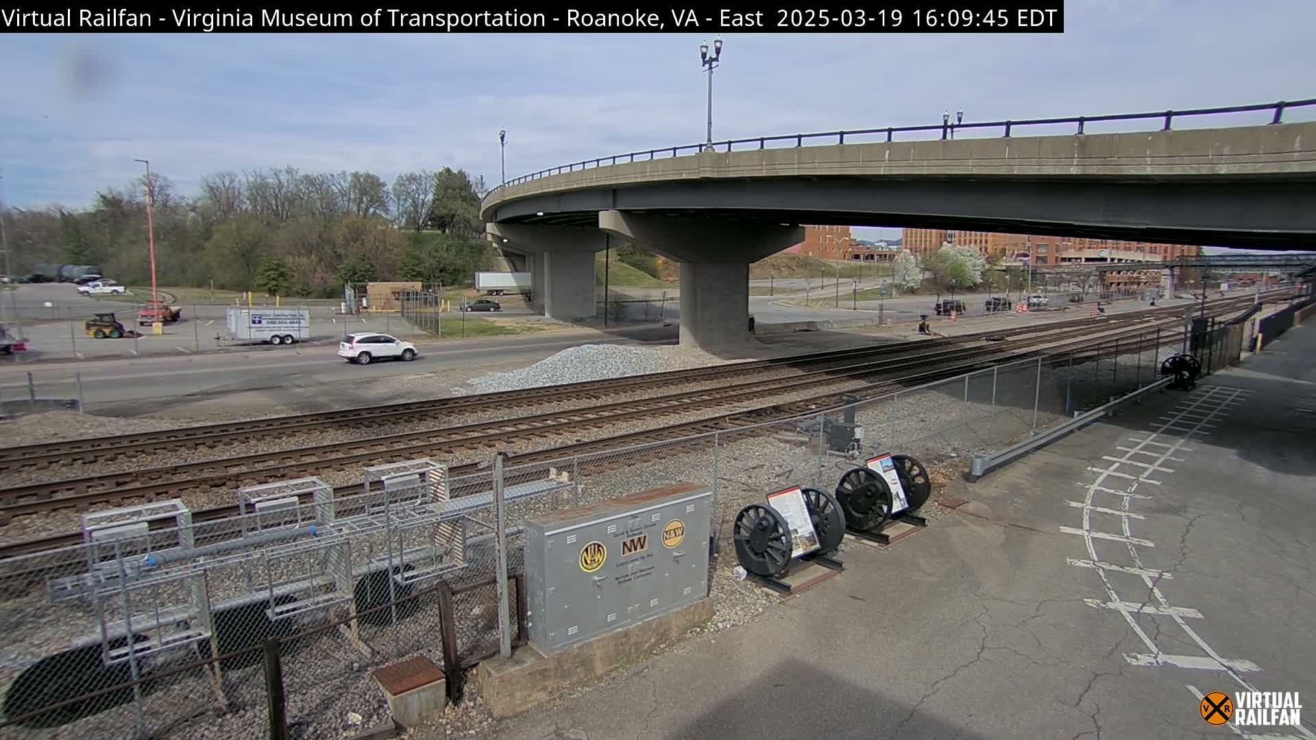 A sunny day reveals a train track system under a highway overpass, with several vehicles visible in a parking lot beyond.