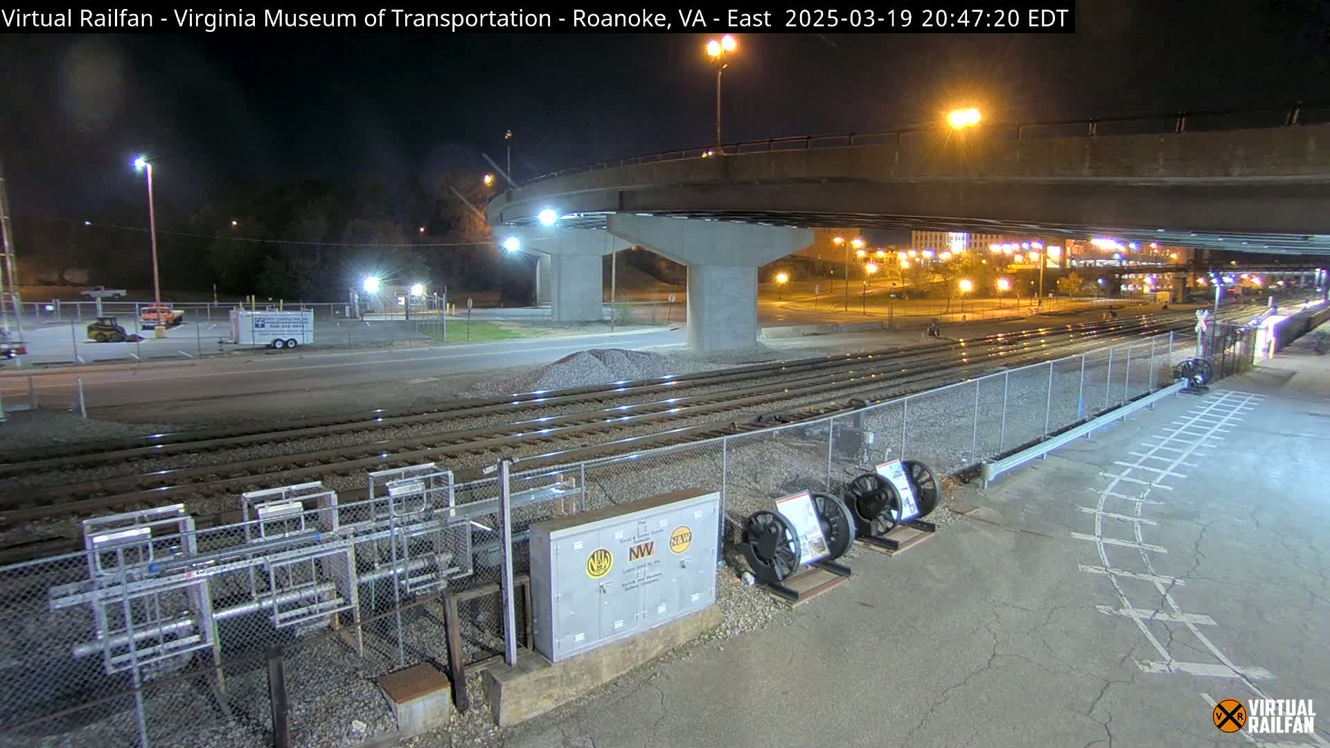 At night, a view of railroad tracks extends under a highway overpass, with several pieces of railway equipment visible nearby.
