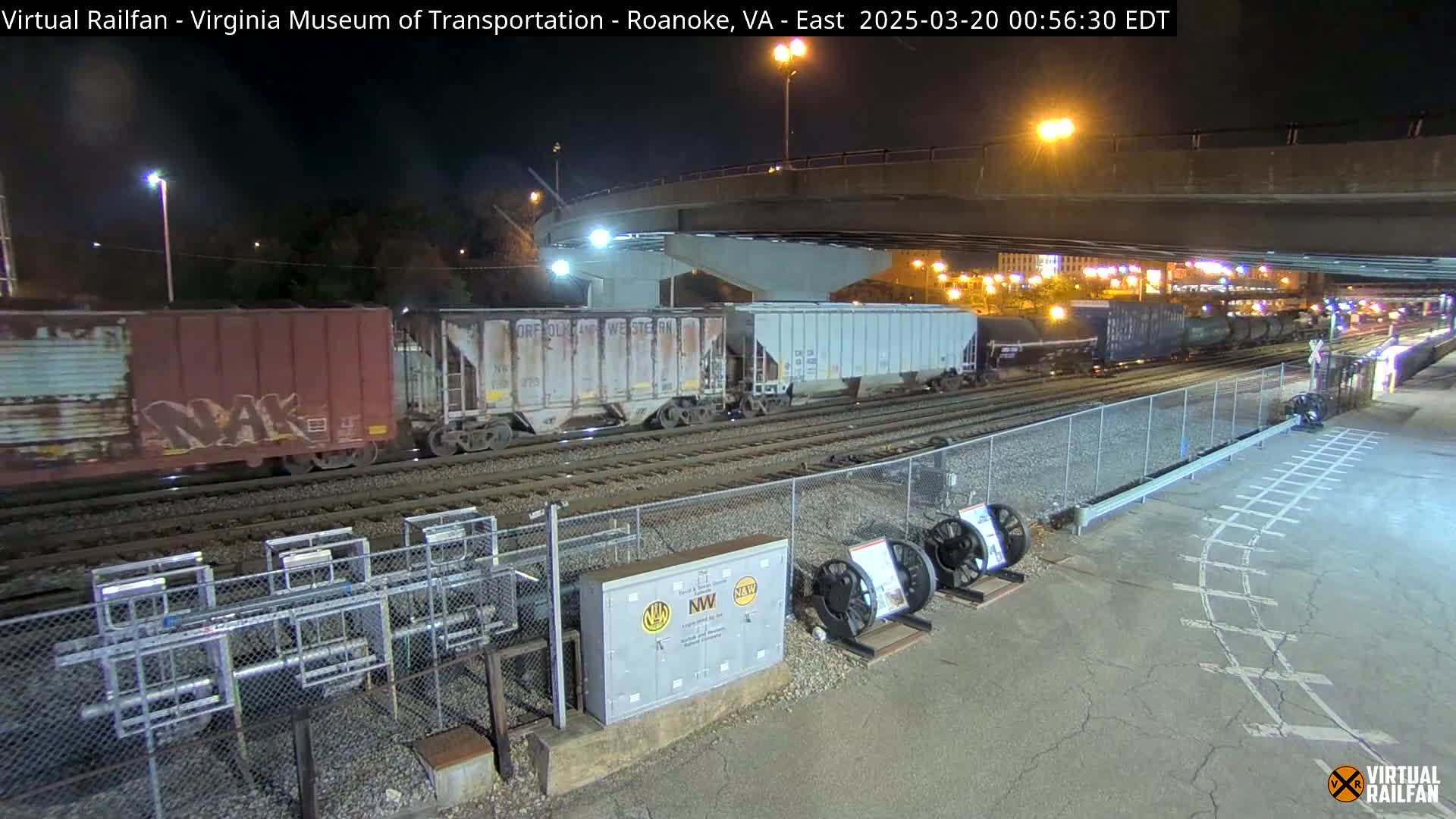 At night, under a highway overpass, a freight train with several boxcars and a few tank cars is parked on railroad tracks next to a fenced area with some equipment and what appears to be old train wheels.