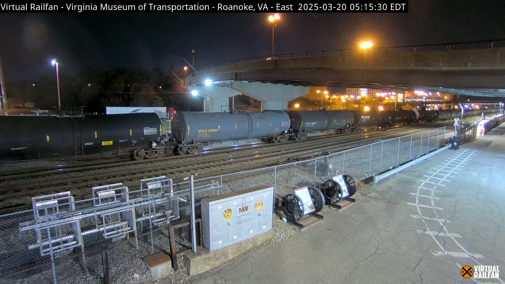 At night, a long train of dark tanker cars sits on railroad tracks beneath a highway overpass.