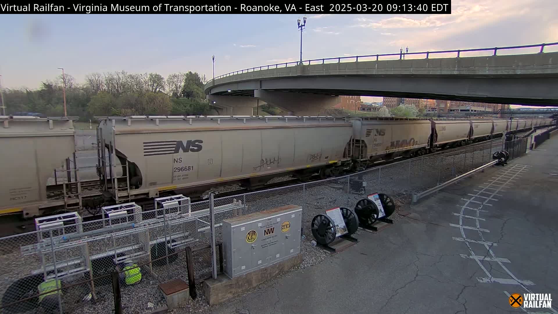 A long freight train of light-grey cars sits under a bridge on a mostly clear day.