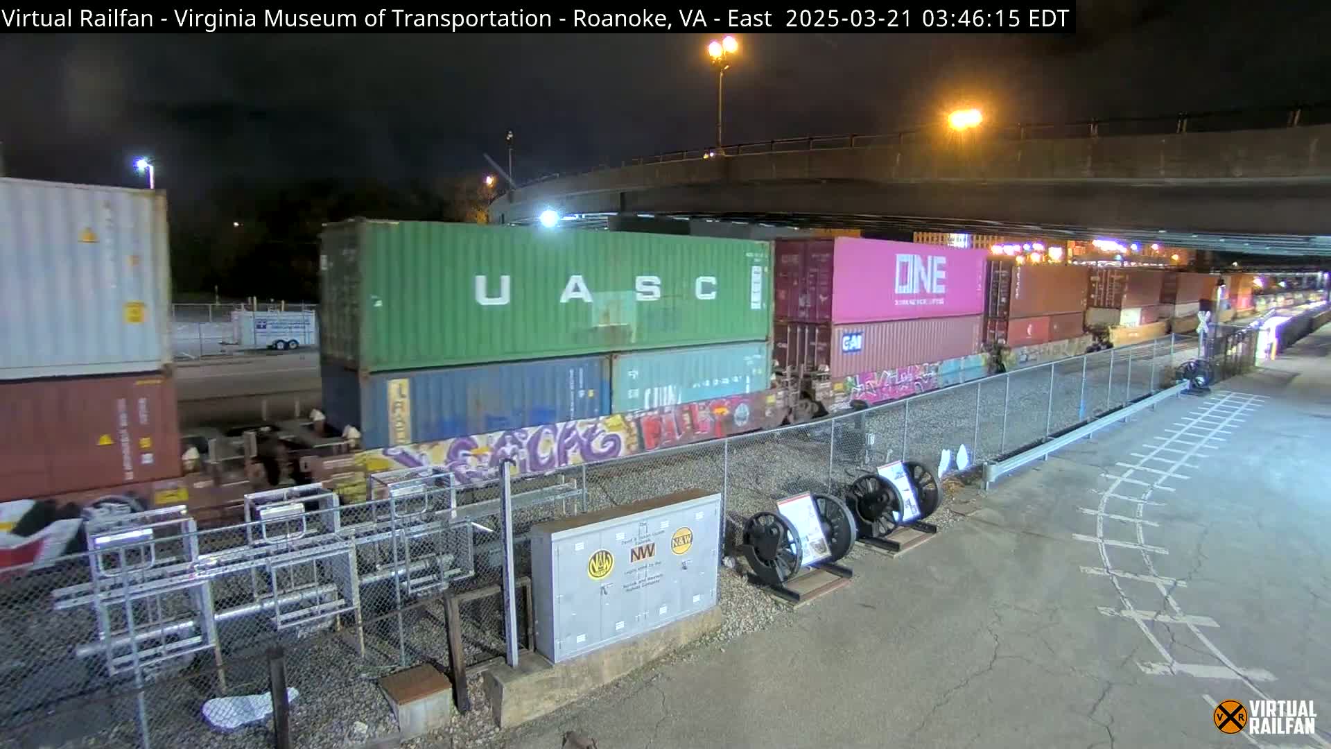 At night, a freight train carrying stacked shipping containers passes under a bridge.