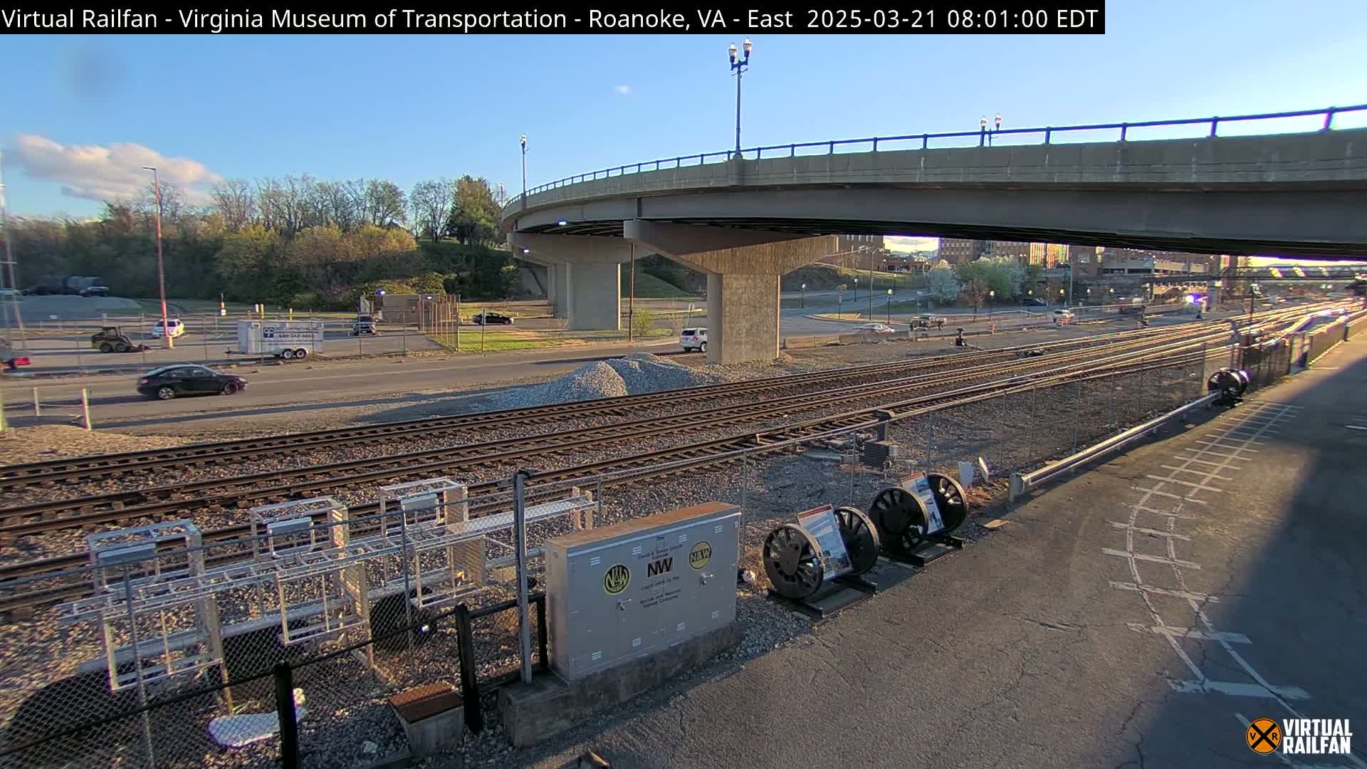 Under a mostly clear sky, a view of railroad tracks and a road runs underneath a bridge, with several cars visible on the road and some construction equipment nearby.