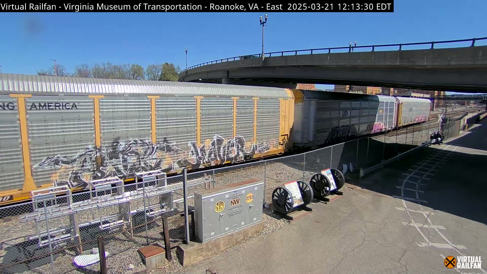 A long freight train with graffiti on its cars sits under a highway overpass on a sunny day.