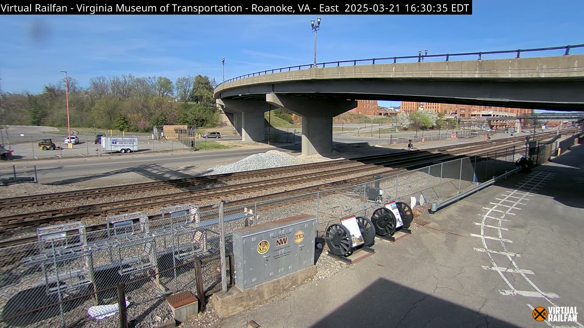 A sunny day reveals a railroad yard with a highway overpass, several parked vehicles, and some railway equipment.