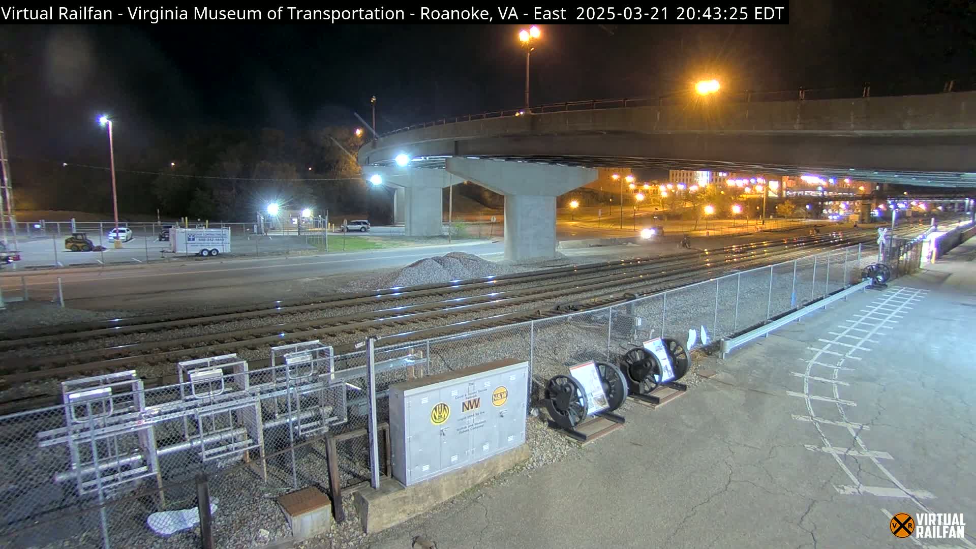 At night, under a bridge, railroad tracks extend toward a road and a parking lot with a few vehicles.