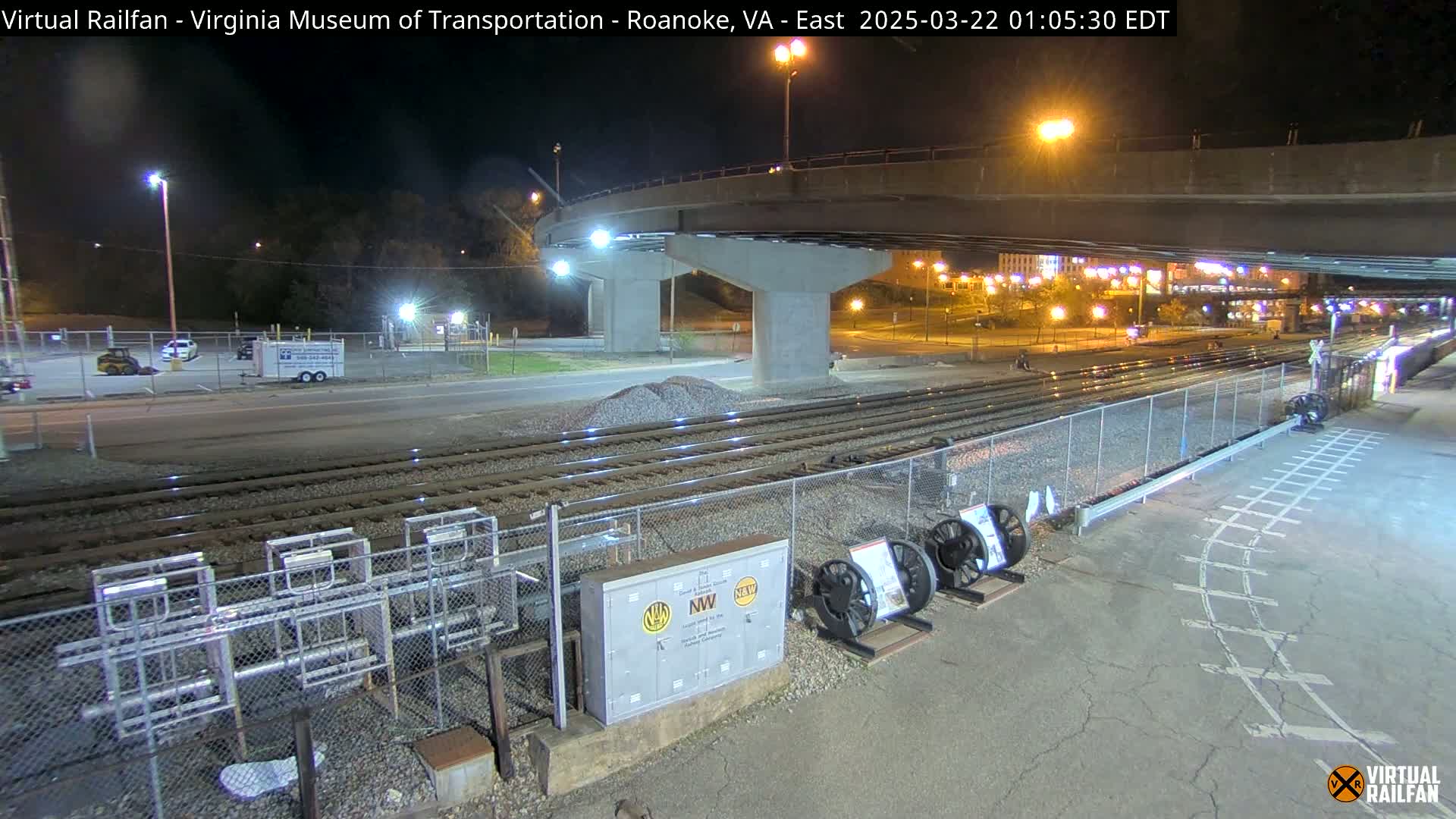 A nighttime view of railroad tracks next to a road, under a highway overpass, with some parked vehicles and equipment visible in the foreground.