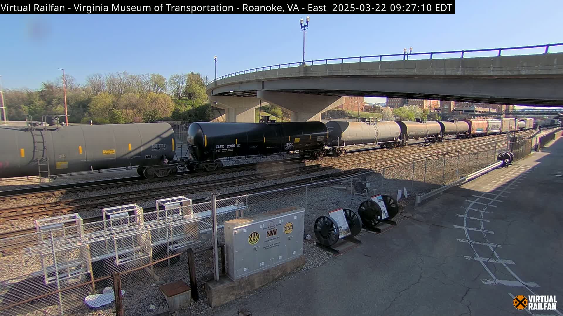 A long freight train of tanker cars passes under a bridge on a sunny day.