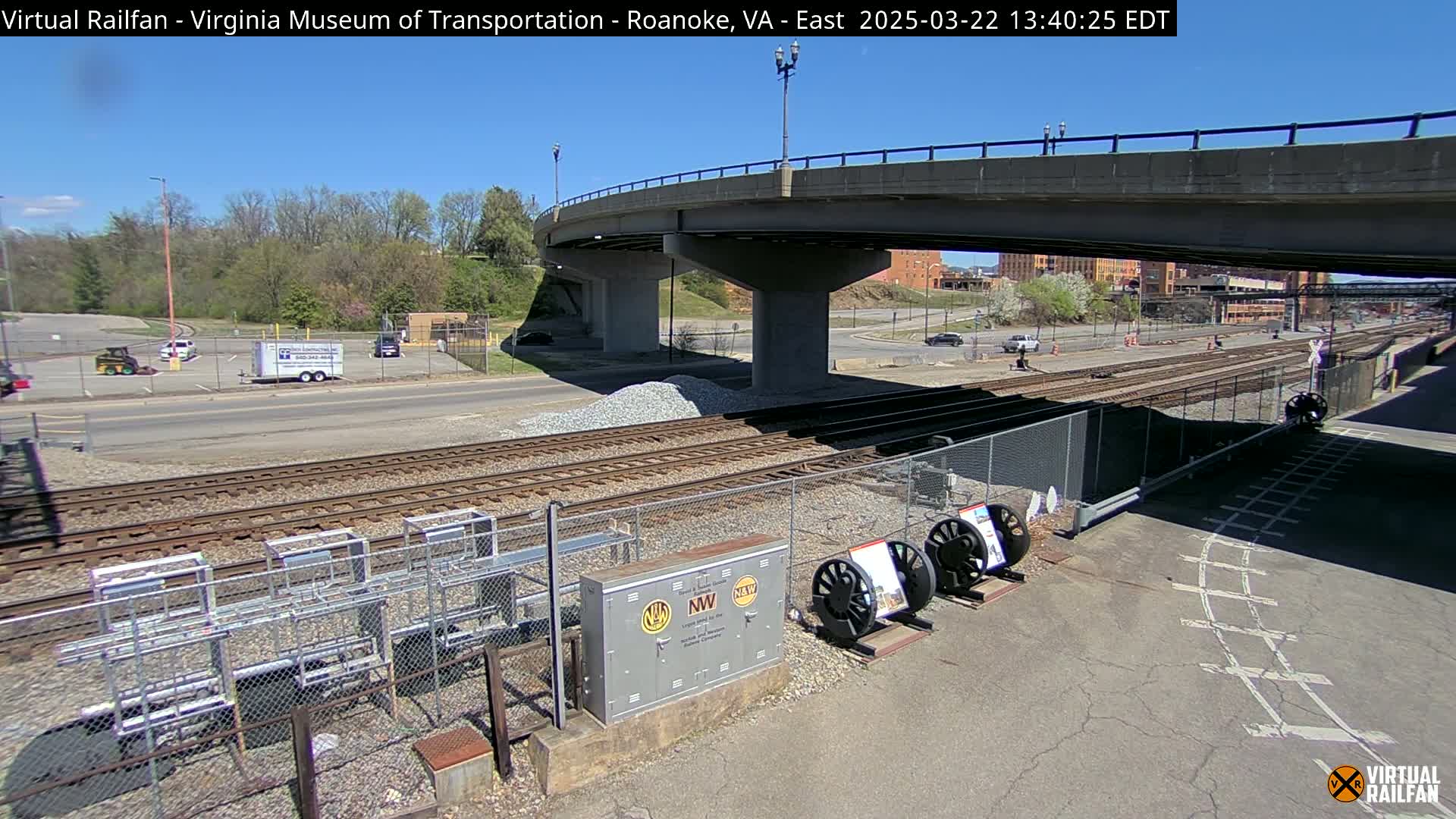 A sunny day reveals railroad tracks, a road, a bridge, and several parked vehicles near a building under a partly cloudy sky.
