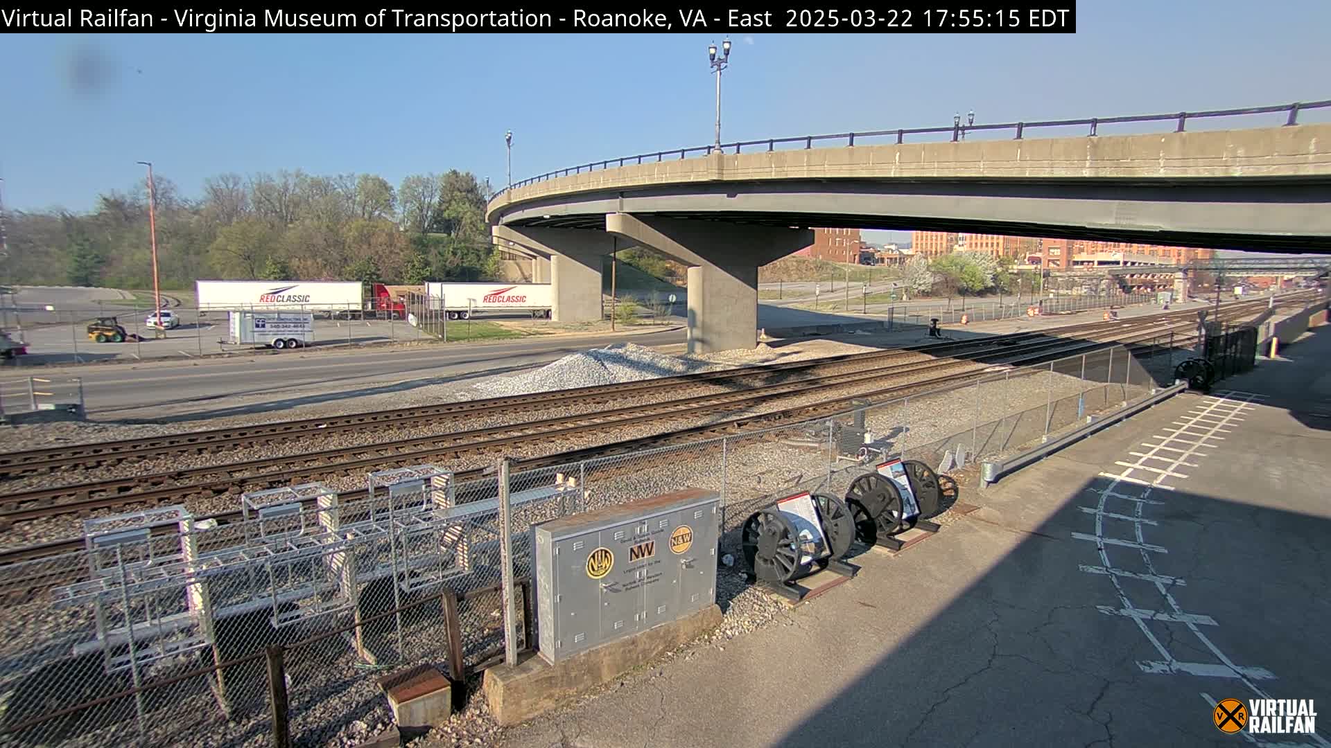A sunny day shows a view of railroad tracks next to a road under a bridge, with several semi-trucks parked nearby and some railway equipment displayed on the ground.