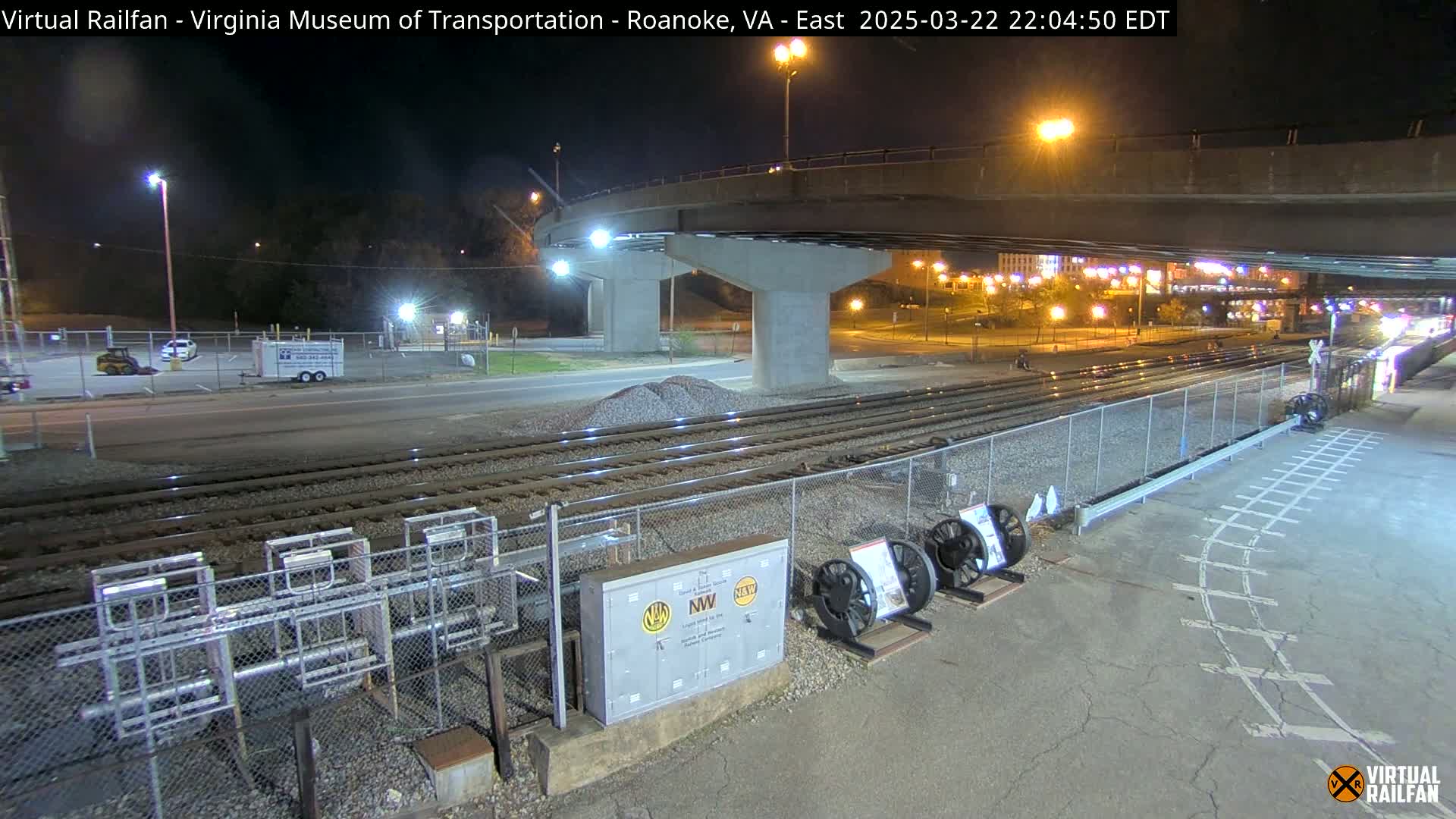 A nighttime view of railroad tracks and a road passing under a bridge, with some equipment and vehicles visible in a parking lot.