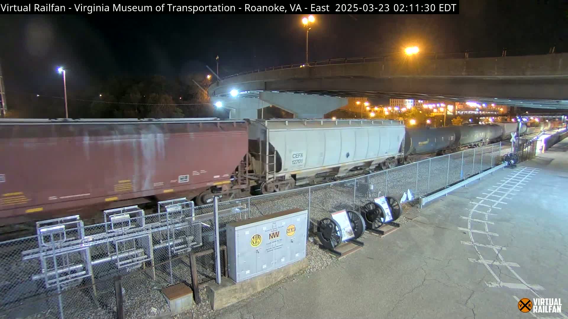 A freight train passes under a highway overpass at night.