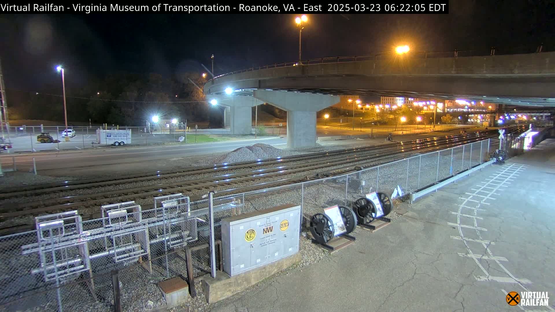 Under a nighttime sky, railroad tracks extend beneath a bridge and past a fenced area with equipment, adjacent to a roadway and parking lot.