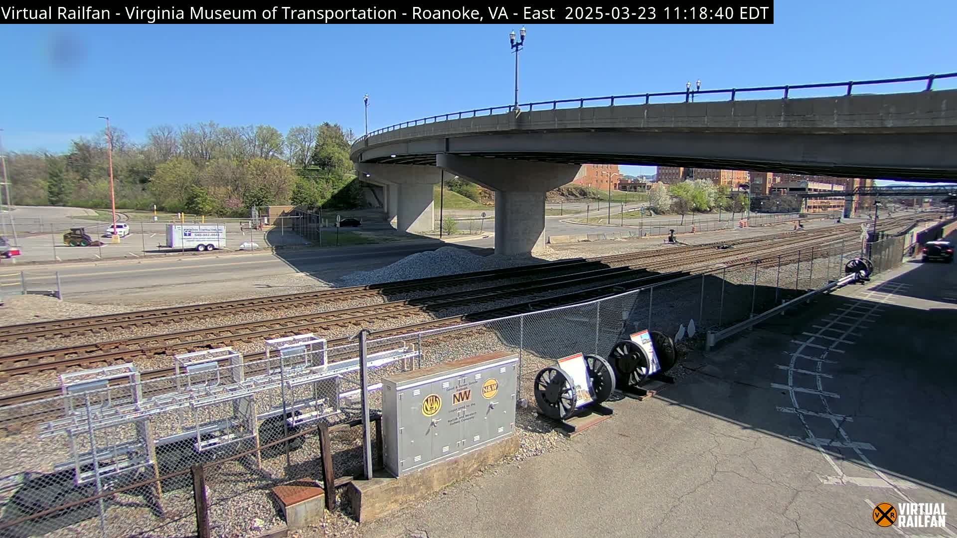 Under a clear blue sky, a roadway overpass crosses over railroad tracks, with some construction equipment and railway components visible below.