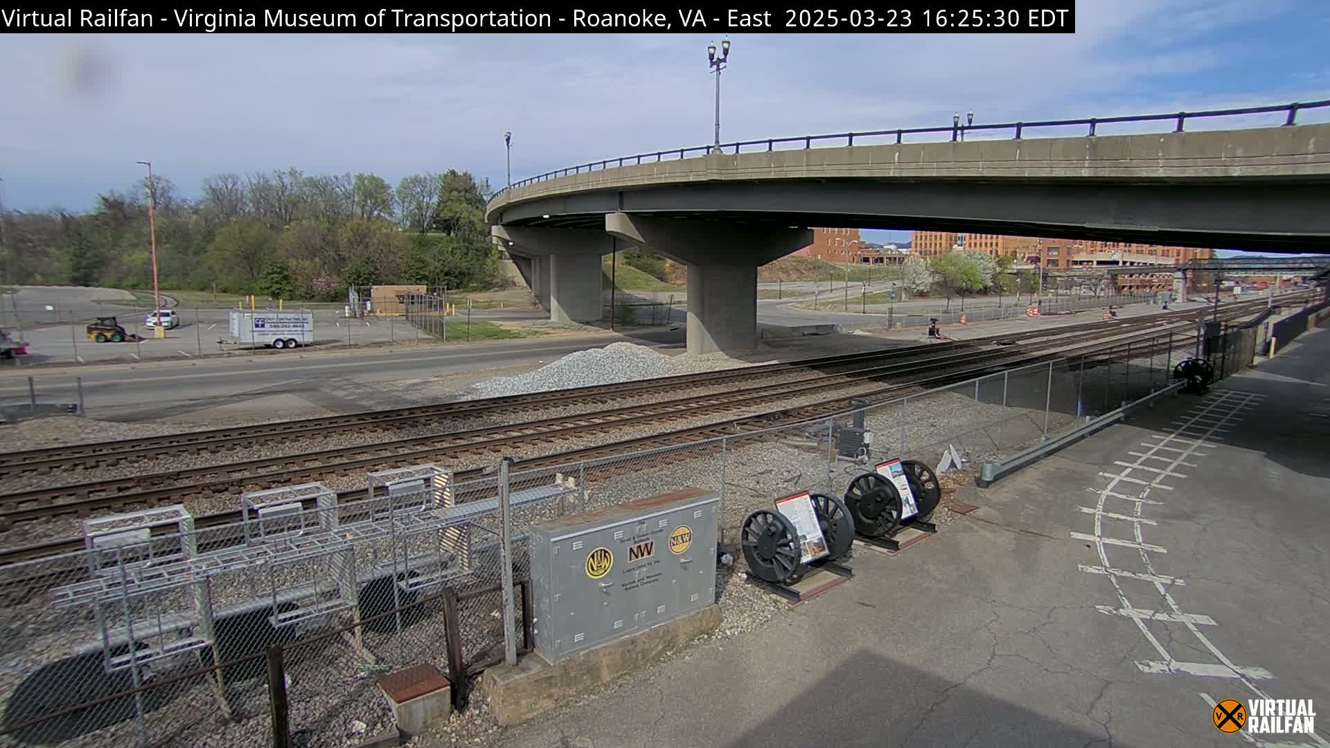 A view of railroad tracks under a bridge on a partly sunny day, with a road and parking lot visible in the background.