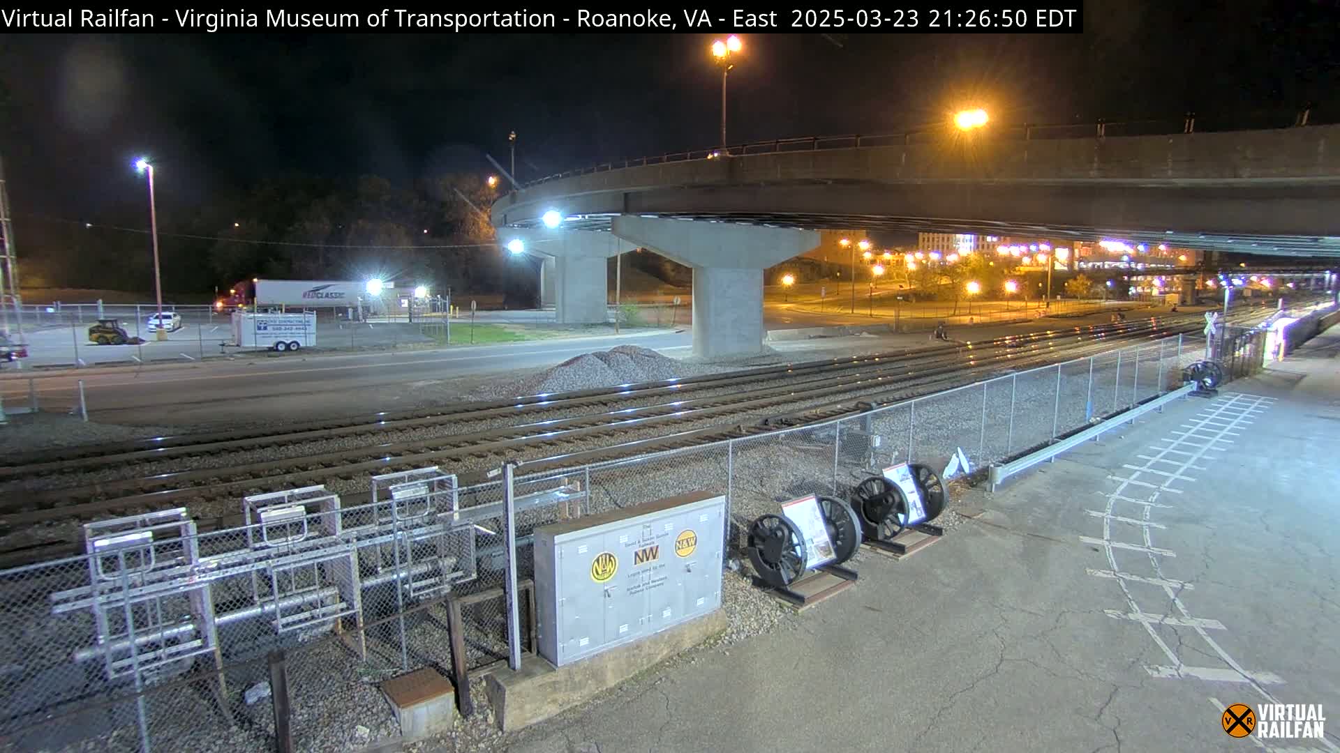 At night, under a bridge, railroad tracks extend toward a roadway and a parking lot with a few vehicles; some equipment is visible near the tracks.