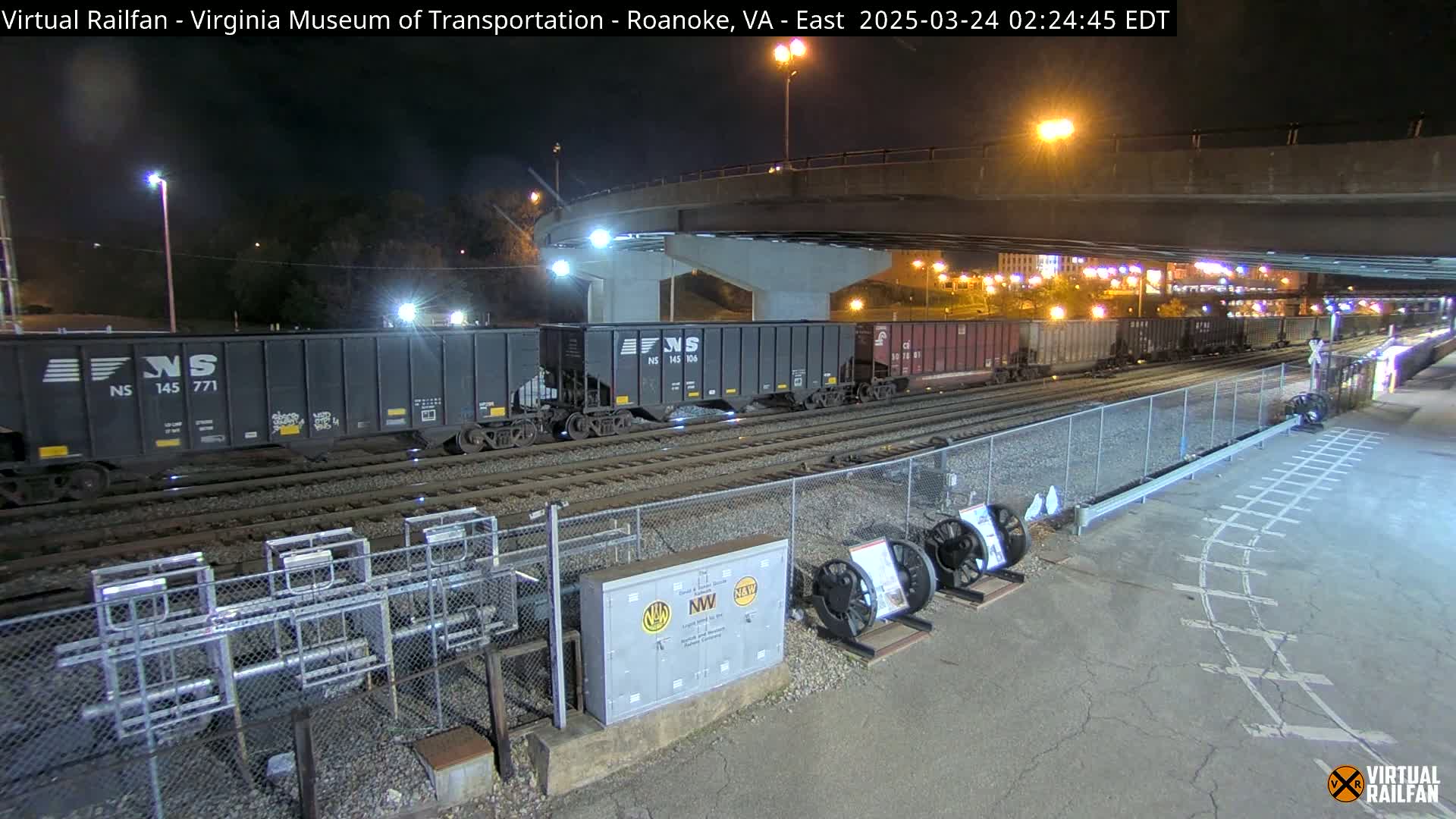 At night, several freight cars sit on railroad tracks under a bridge, with additional cars visible in the distance.