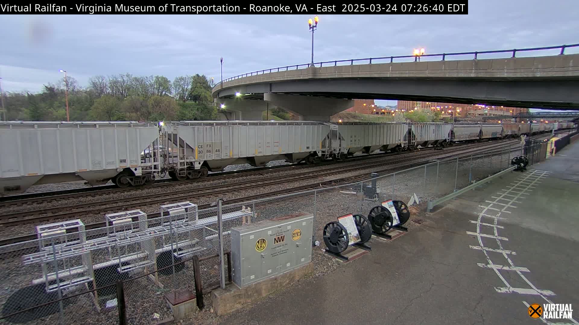 A long freight train of gray covered hopper cars passes under a bridge on an overcast day.