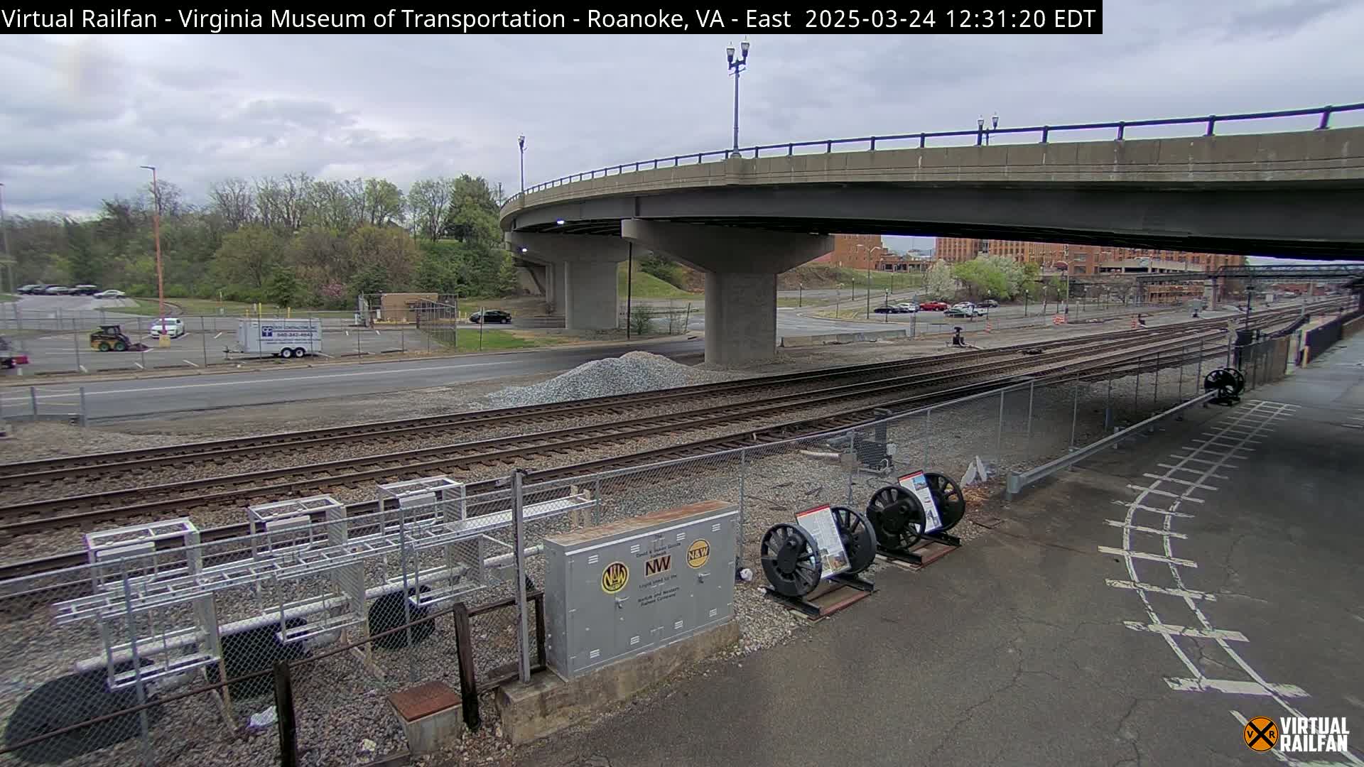 A partly cloudy day shows a view of railroad tracks beneath a highway overpass, with several vehicles parked nearby and some construction materials in a pile.