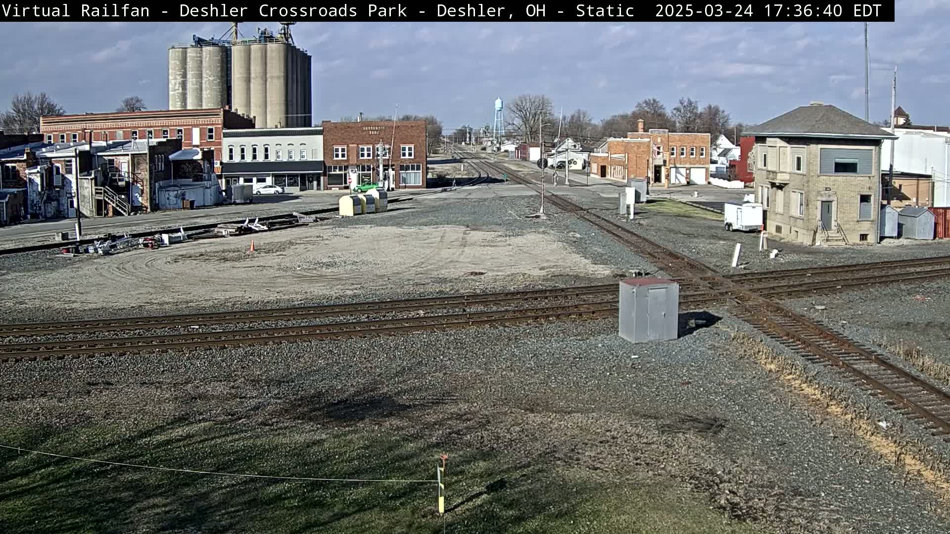 A sunny day reveals a railroad crossing in a small town, with buildings, grain silos, and train tracks visible.