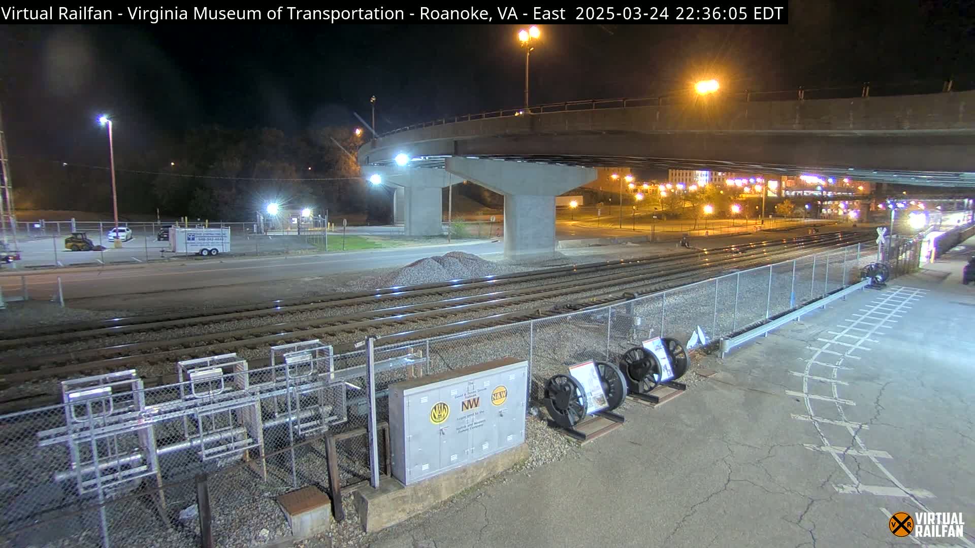 At night, under a highway overpass, railroad tracks extend toward a distant cityscape, with several pieces of railway equipment visible nearby.