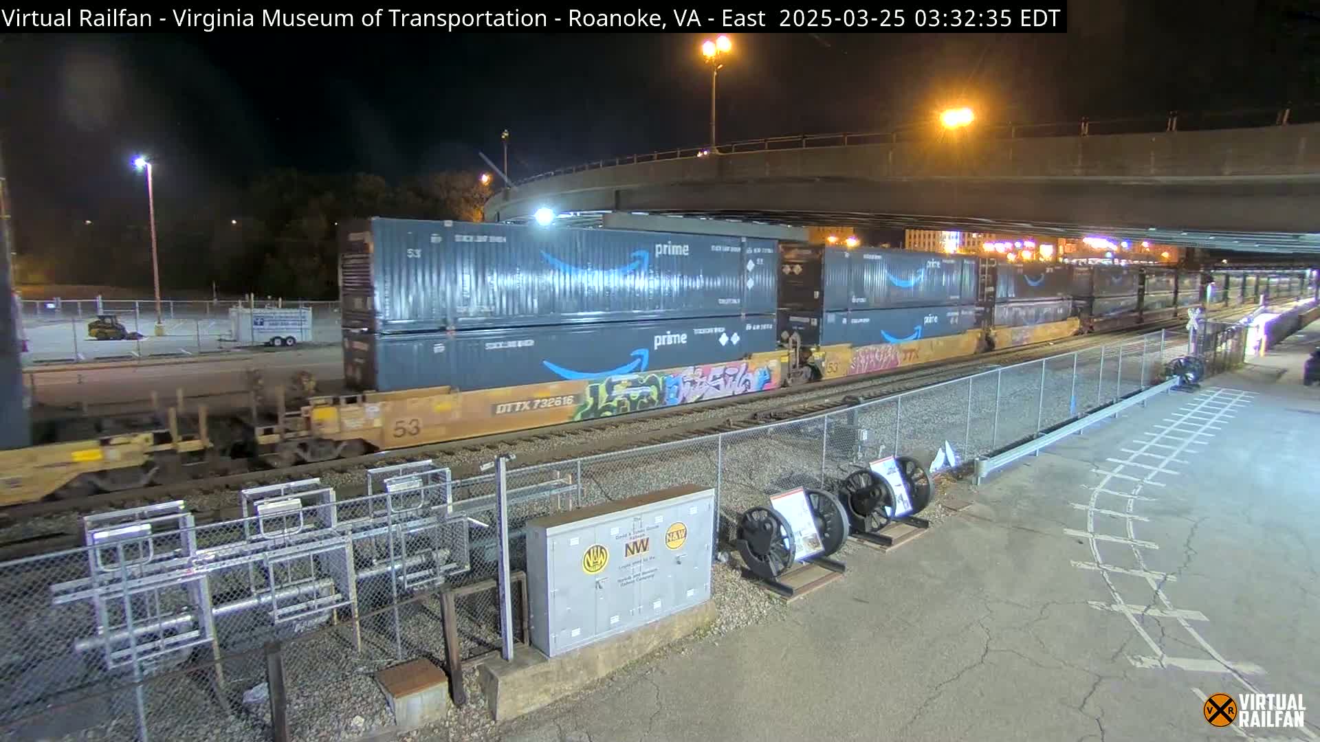 At night, a freight train carrying several Amazon Prime containers passes under a highway overpass.