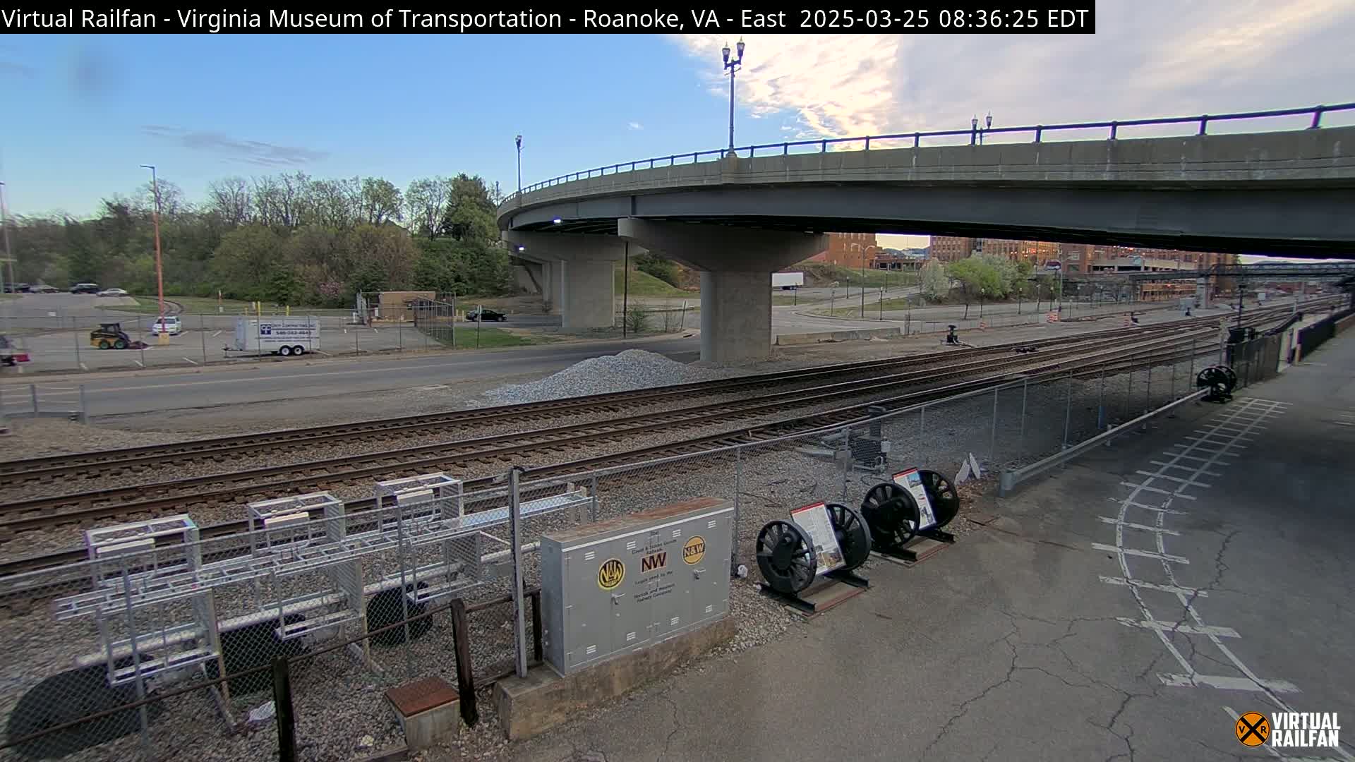A clear, daytime view of railroad tracks under a highway overpass, with some construction equipment and vehicles visible in a parking lot beyond.