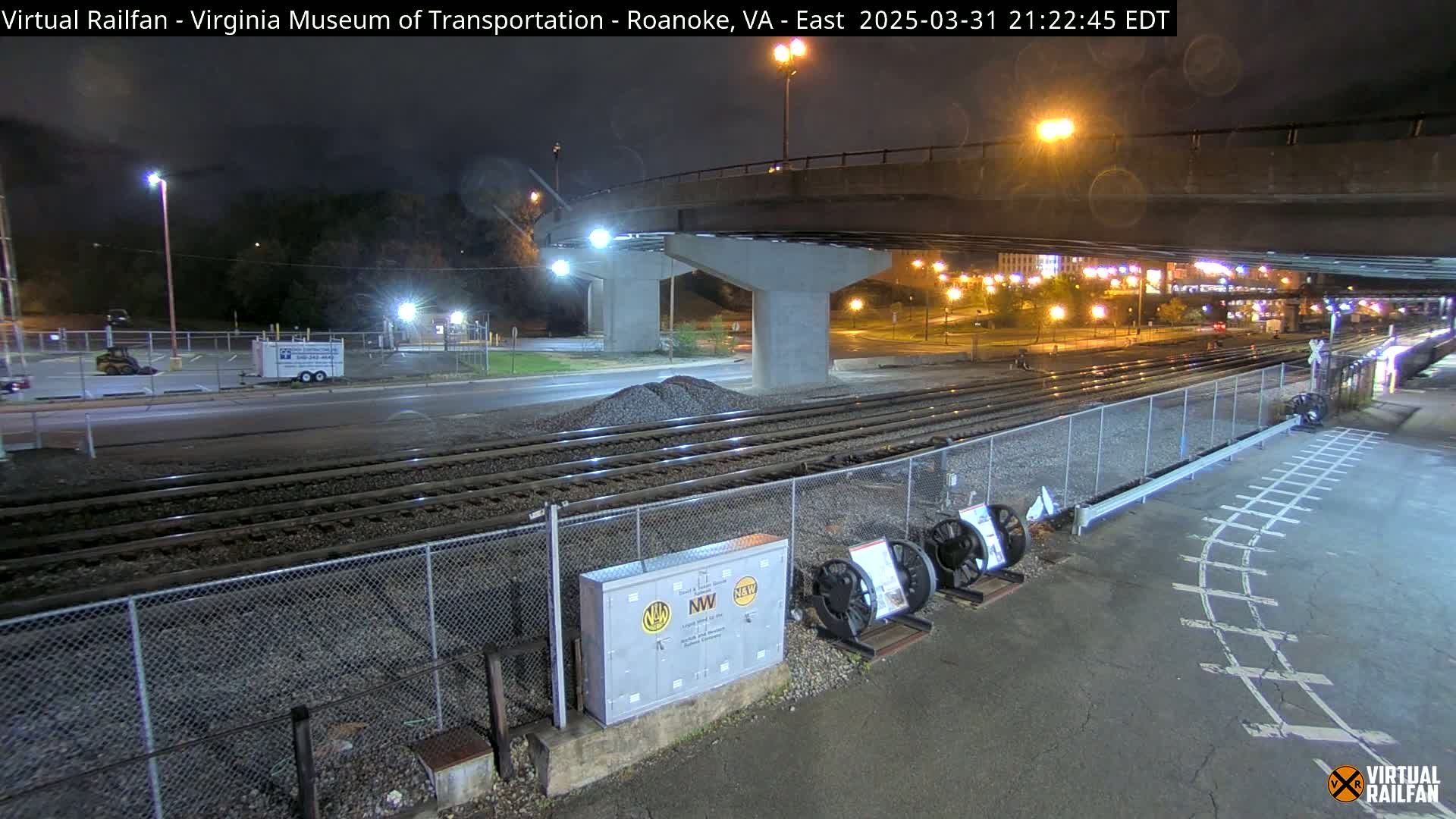 A nighttime view of railroad tracks under a bridge, with a road and some parked vehicles visible in the background.