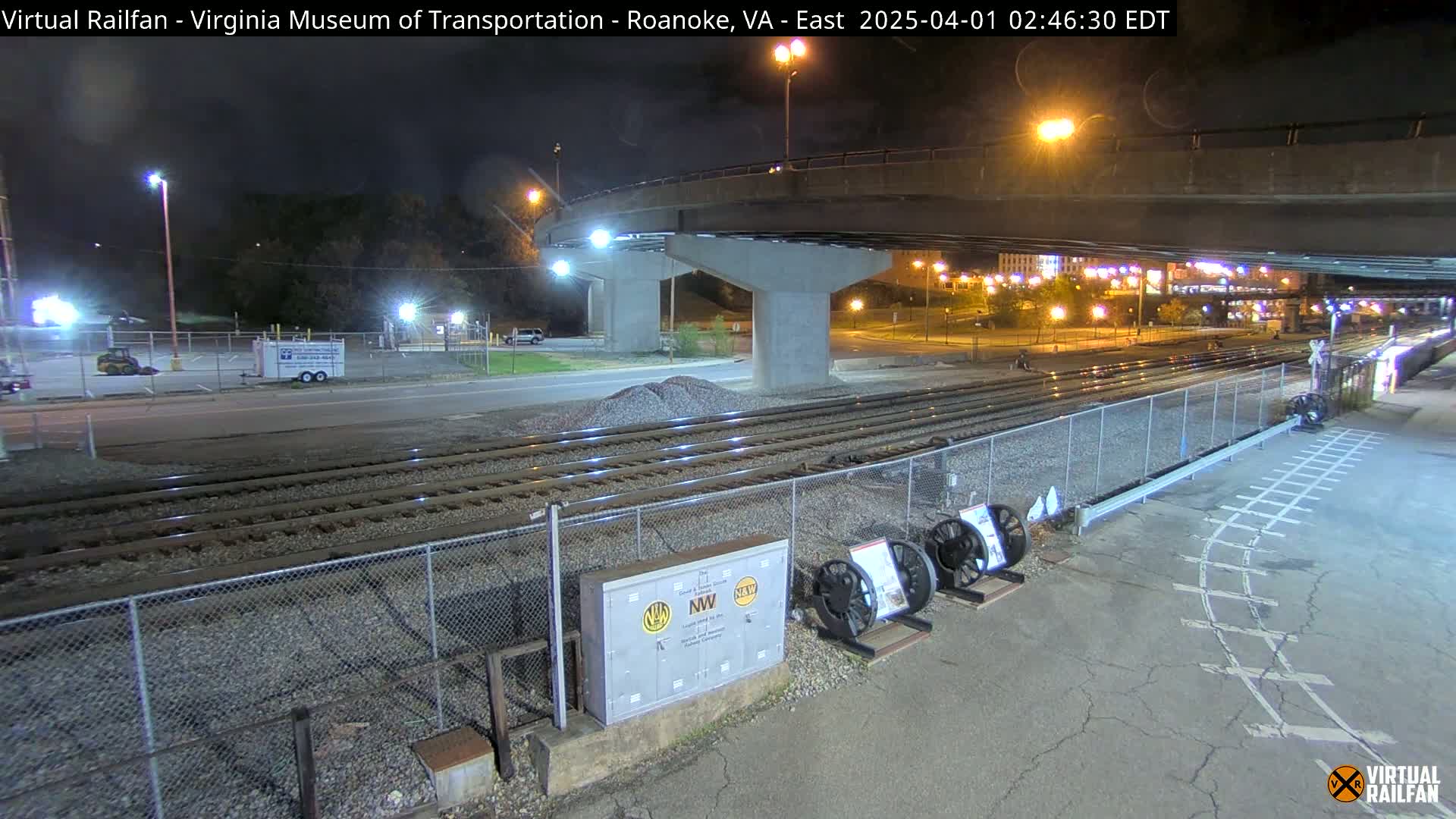 At night, under a bridge, train tracks extend into the distance, past a chain-link fence and some equipment.