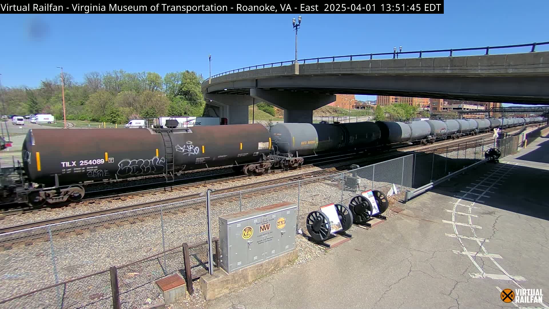 Under a sunny sky, a long freight train of tanker cars passes under a bridge near a chain-link fence.