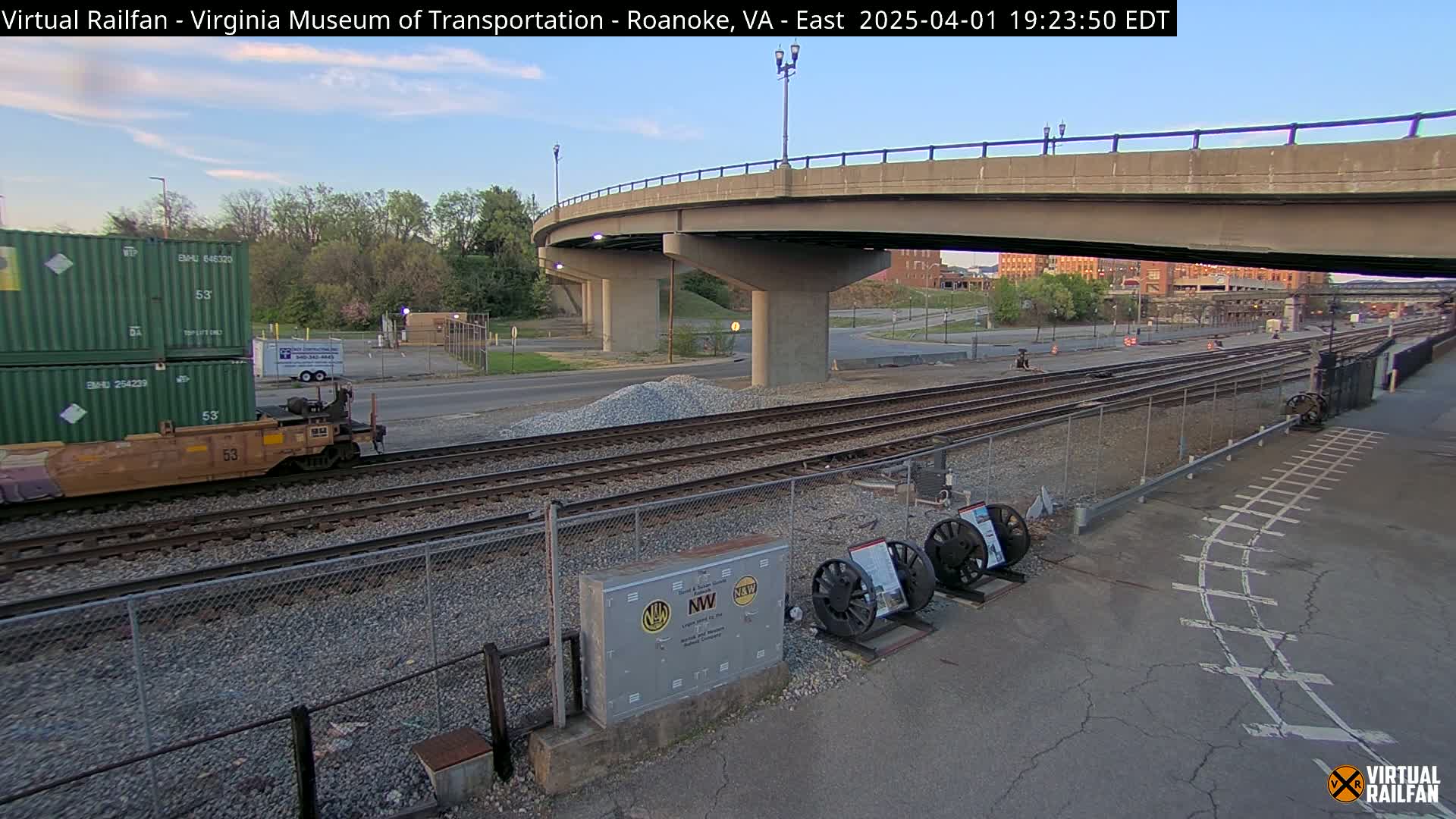 A train car carrying two stacked green shipping containers sits on railroad tracks under a highway overpass at dusk.